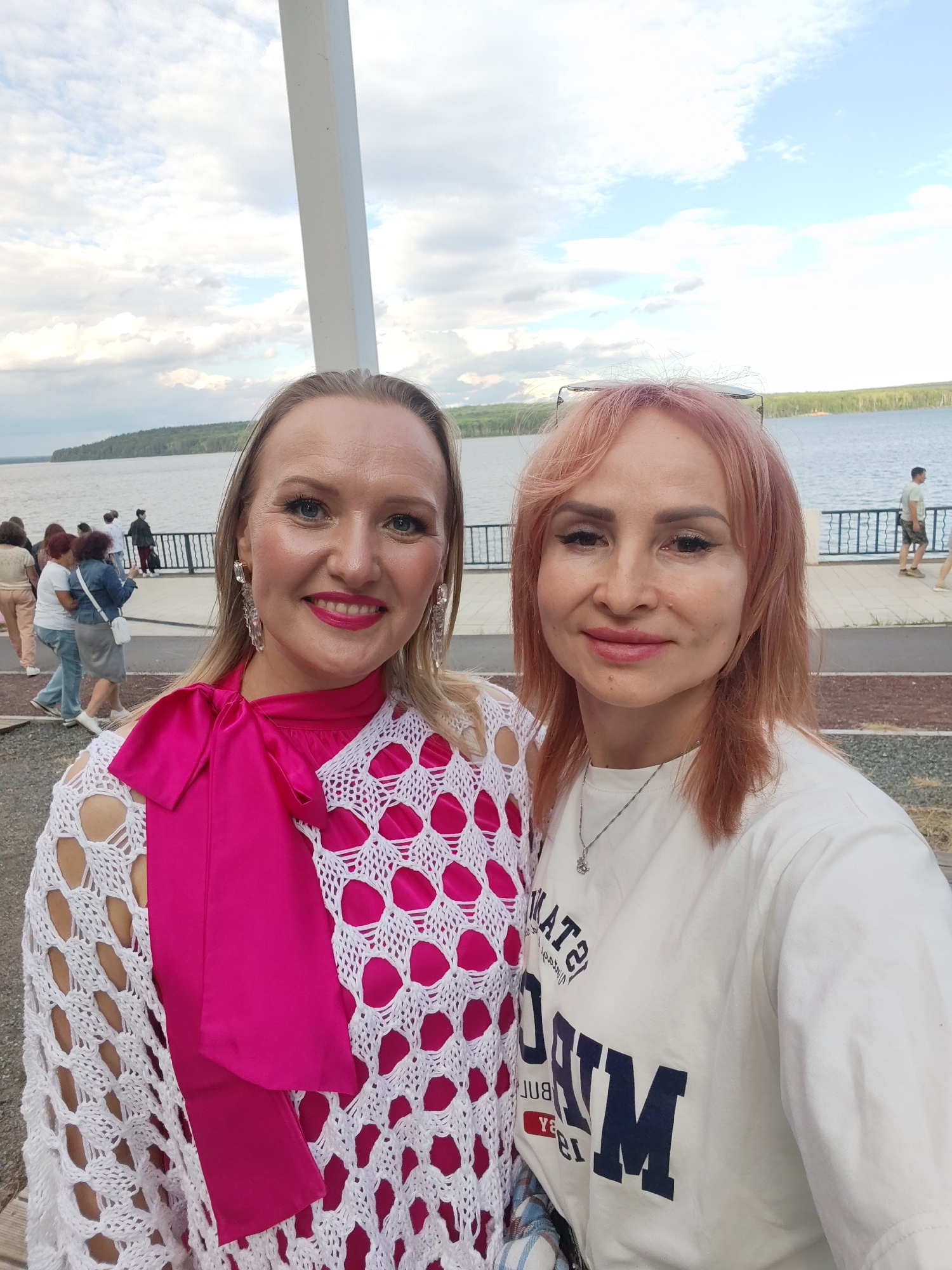 Two women are taking a selfie outdoors with a body of water and a treeline in the background. The woman on the left has blonde hair and is wearing a bright pink dress with a white crocheted shawl and large dangling earrings. She has bright pink lipstick and is smiling. The woman on the right has reddish-blonde hair and is wearing a white t-shirt with dark blue lettering. She is also smiling. The sky is cloudy, suggesting it is either morning or late afternoon. The scene appears to be a public walkway along the water, with a railing and pavement visible. There are a few other people walking in the background. The overall mood is cheerful and relaxed. The text on the woman's t-shirt appears to be in a varsity font, though it is partially obscured.