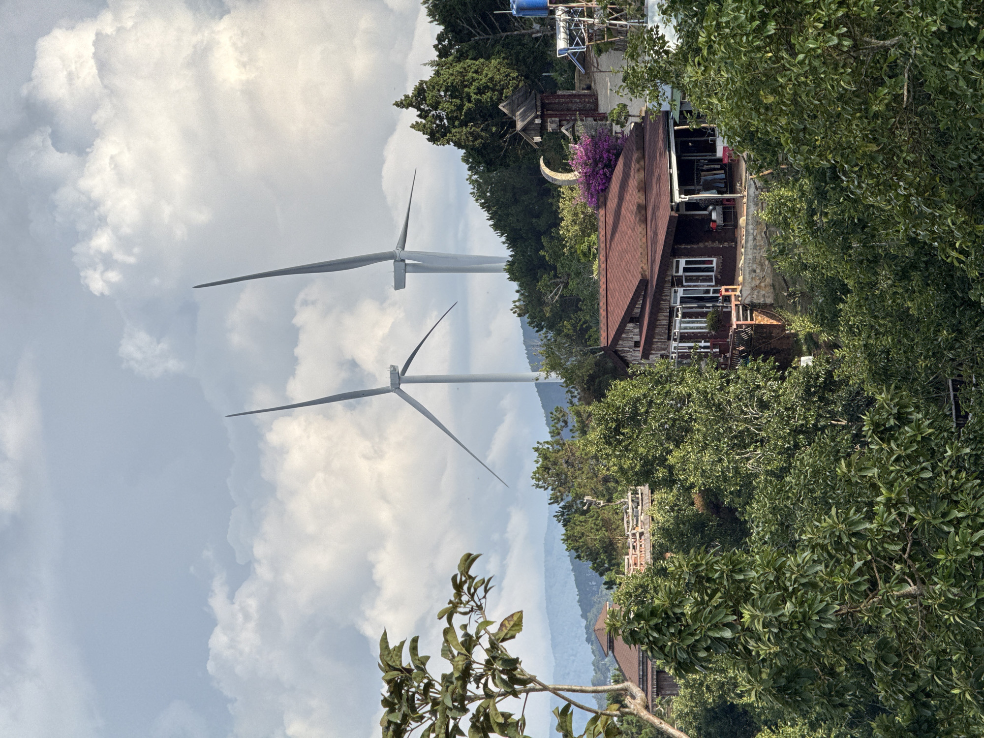 The image shows two wind turbines against a cloudy sky. Adjacent to the turbines is a cluster of trees and a wooden house with a brown roof. The location is Phát Chi, Vietnam.