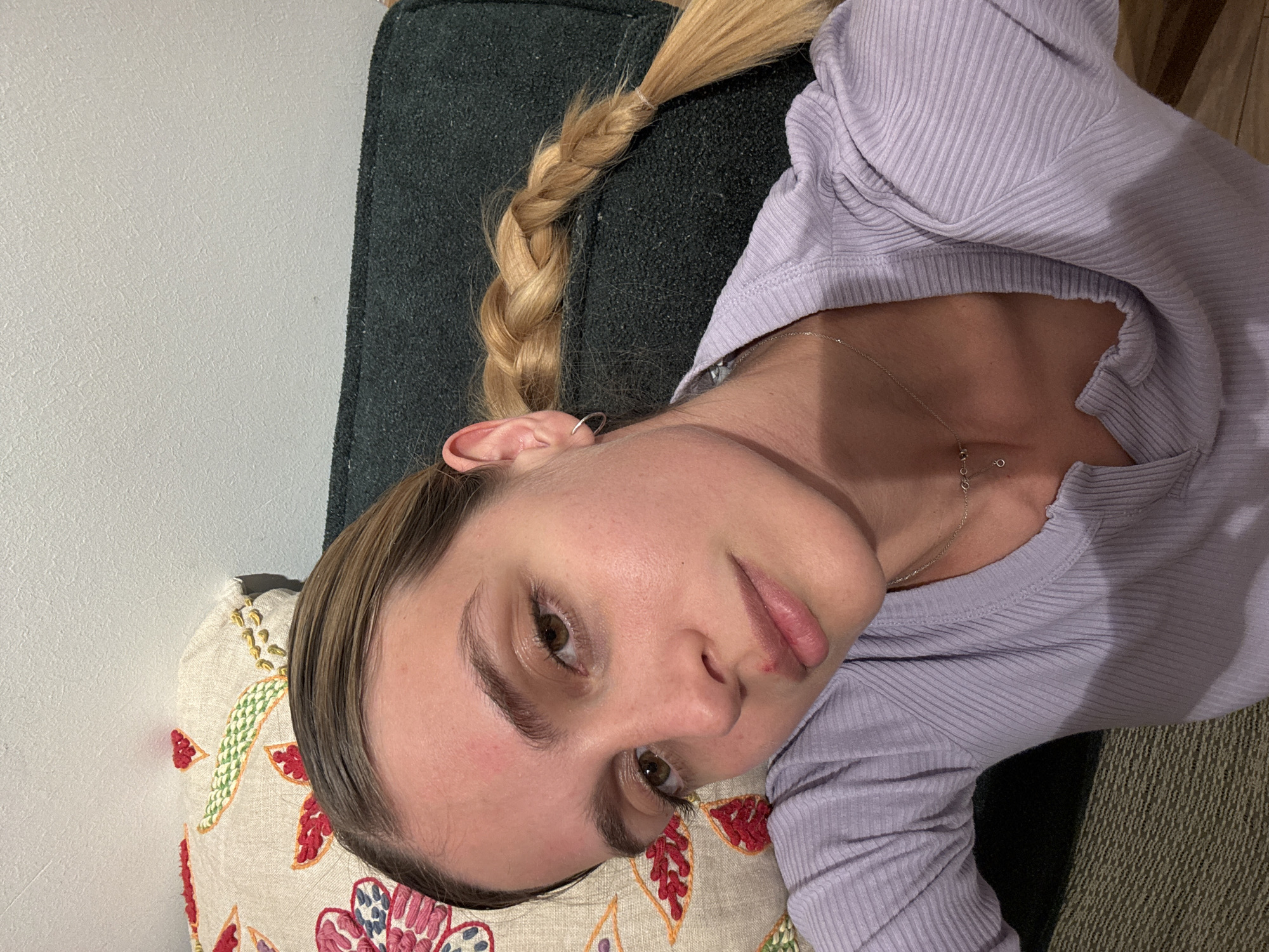 A young person, likely female, is depicted reclining indoors, looking directly at the camera. Their head is resting on a decorative pillow, and their body is positioned on what appears to be a dark, textured sofa or chair.
The subject has light skin, brown eyes, and light brown or blonde hair styled in a long braid that rests over their shoulder. They are wearing a light purple, long-sleeved, ribbed top with a scoop neck. A delicate silver chain necklace with a small charm hangs around their neck. A small silver hoop earring is visible in their left ear. Their expression is neutral and calm, with their lips slightly parted, revealing a minor redness or chapping on the lower lip.
The pillow beneath their head is made of cream-colored fabric and features colorful embroidered patterns of red, green, orange, and blue floral or leaf designs. The background shows a light-colored, textured wall. The lighting suggests an indoor setting, possibly with artificial light, creating soft shadows. There is no visible text in the image.