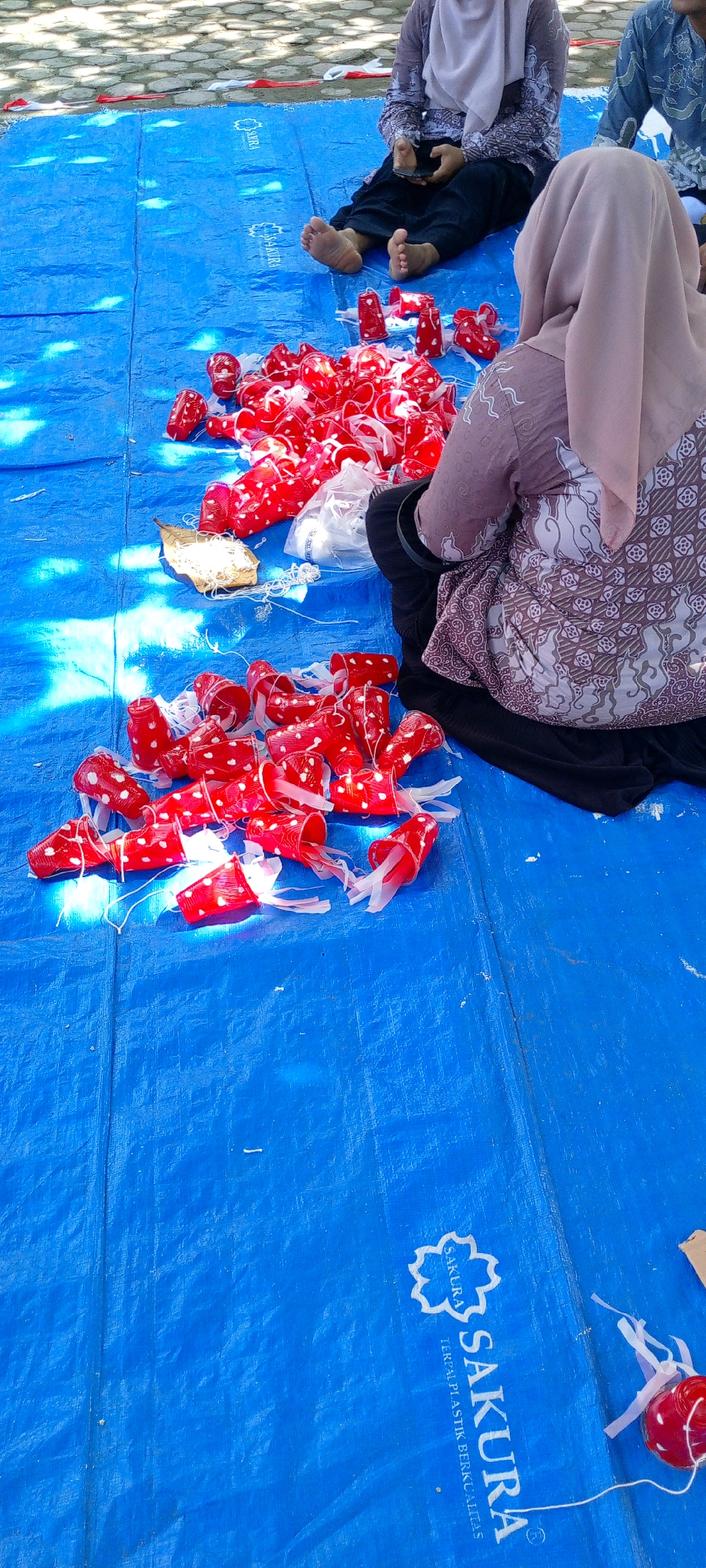 In Baratpayaitek, Indonesia, a group of people are gathered on a blue tarp, engaged in a craft activity. The scene appears to be outdoors, with dappled sunlight suggesting the presence of trees. The focus is on a collection of small, red, mushroom-shaped objects, each adorned with white polka dots and attached to white string. These objects are scattered across the tarp, indicating they are either being assembled or prepared for use. Two individuals are visible, both wearing headscarves and traditional Indonesian clothing. One person, seated with their feet bare, is holding a mobile phone. Another individual, also seated, is partially visible from behind, wearing a patterned headscarf and clothing. The blue tarp itself features a repeating "SAKURA" logo. The overall impression is one of a community event or a workshop focused on creating decorative items.