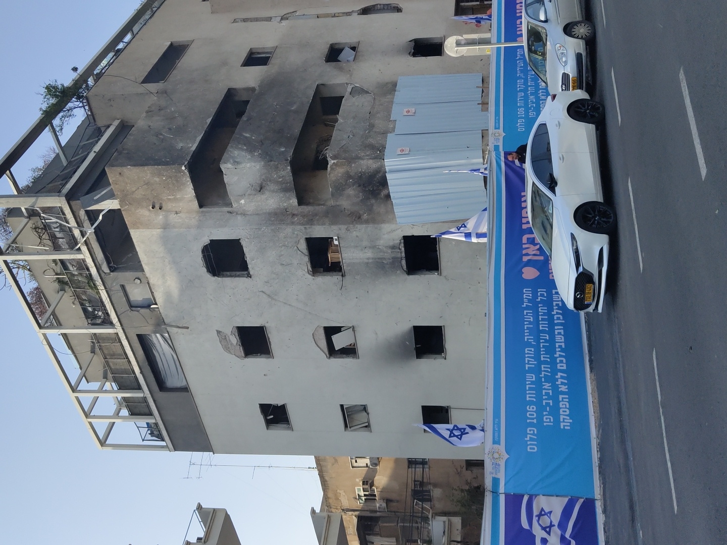The image captures an urban street scene in Tel-Aviv, Israel, during daytime under a clear blue sky. The most prominent subject is a multi-story building that appears significantly damaged. Its facade is marked with black charring and soot, and numerous windows are shattered or completely missing, suggesting it may have been affected by a fire or other destructive event. Some upper floors show more extensive damage, and metal framework from balconies or structures protrudes. Greenery is visible on some of the higher levels.
Along the street level, a large blue banner stretches horizontally, displaying text in Hebrew and multiple Israeli flags. The text on the banner reads, "City Hall Command Center, Service Hotline 106 Plus, and all units of Tel Aviv-Yafo Municipality, for you (plural) non-stop." This indicates a public information or support initiative.
Two cars are parked parallel to the curb next to the banner: a white Mazda 3 sedan with black wheels and a partially visible silver Fiat Punto hatchback behind it. The white Mazda has an Israeli license plate (732-18-402). A person is partially visible inside the white Mazda. The overall scene conveys a sense of a community responding to or recovering from an impactful event, with visible damage contrasted by public service announcements.