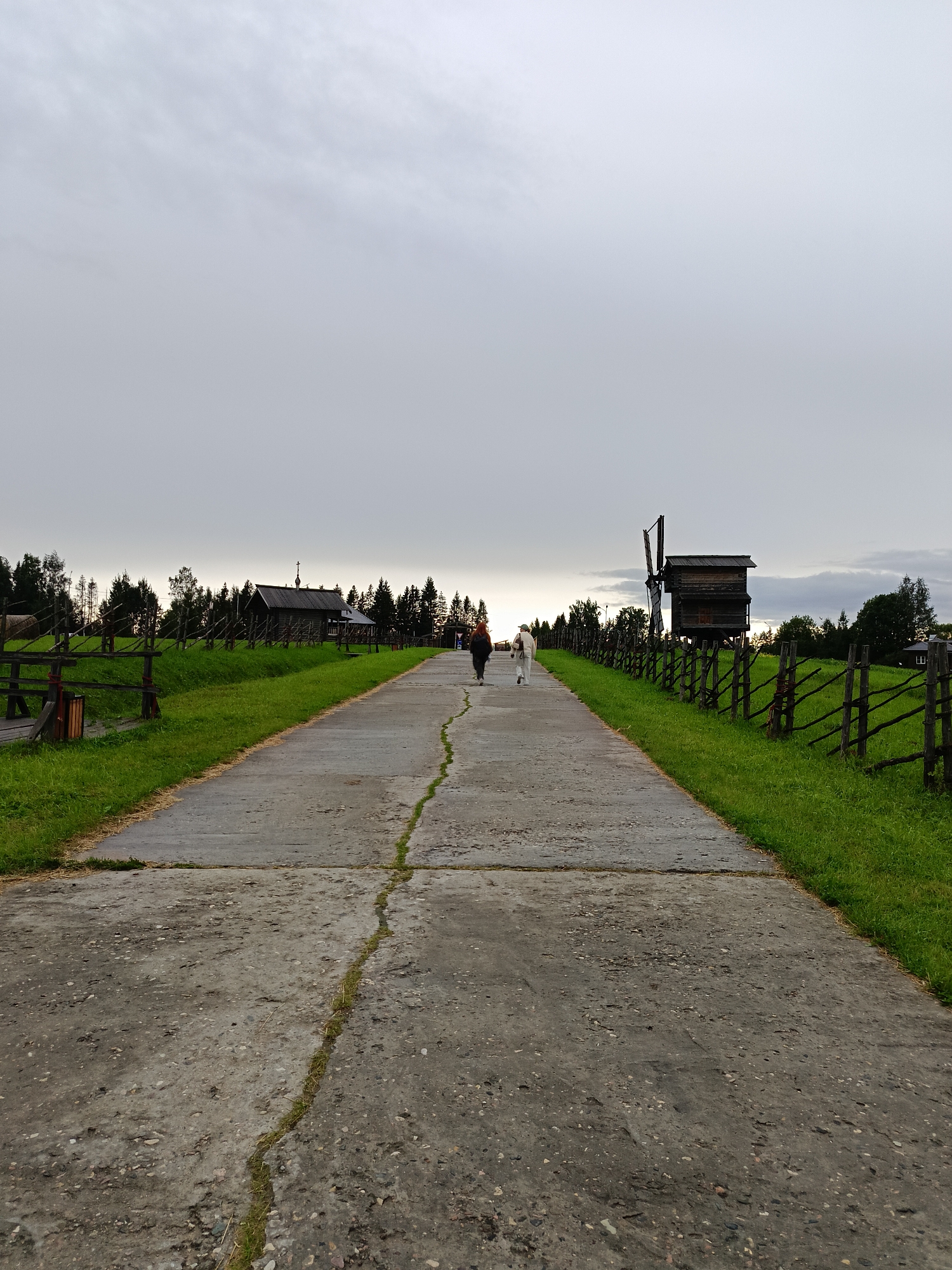 In Semenkovo, Russia, an overcast day frames a scene in an outdoor museum. A concrete path, cracked and overgrown with grass, leads into the distance. On the right, a wooden fence with vertical posts encloses a vibrant green field. Further along the path, a historic wooden windmill with a large, dark propeller stands prominently against the grey sky. To the left, another wooden fence borders a grassy area, and a rustic wooden building with a cross atop its roof is visible. Two individuals are walking away from the camera on the path, one wearing dark clothing and the other in lighter attire. The background is dotted with trees, and a few more wooden structures are sparsely visible. The overall impression is one of a rural, historical setting under a muted sky.