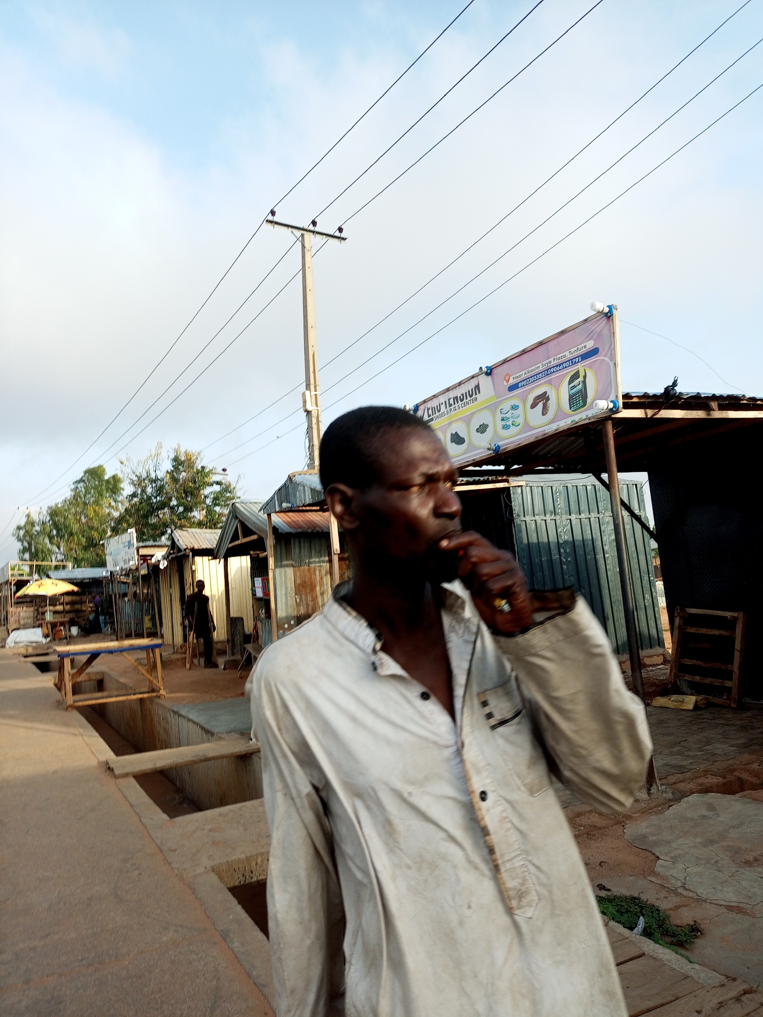 The image depicts a street scene in Garko, Nigeria, under a sky with scattered clouds. In the foreground, a man wearing a light-colored, button-down shirt is the primary subject. He is shown from the chest up, with his face in profile as he brings his hand to his mouth, possibly smoking or chewing something.
Behind the man, a street market is visible. Several stalls with corrugated metal roofs line the dusty path. A prominent banner advertising "CHUO TEN GUN SHOES P.O.S CENTER" is stretched across a pole, featuring images of shoes, a phone, and what appears to be a handgun. Power lines crisscross the sky above the market. In the mid-ground, a man with crutches stands near a stall, and further down the street, other individuals and market activities are suggested by the presence of umbrellas and structures. The ground is a mix of dirt and concrete with some open drainage channels. The overall impression is that of a bustling, open-air market environment.