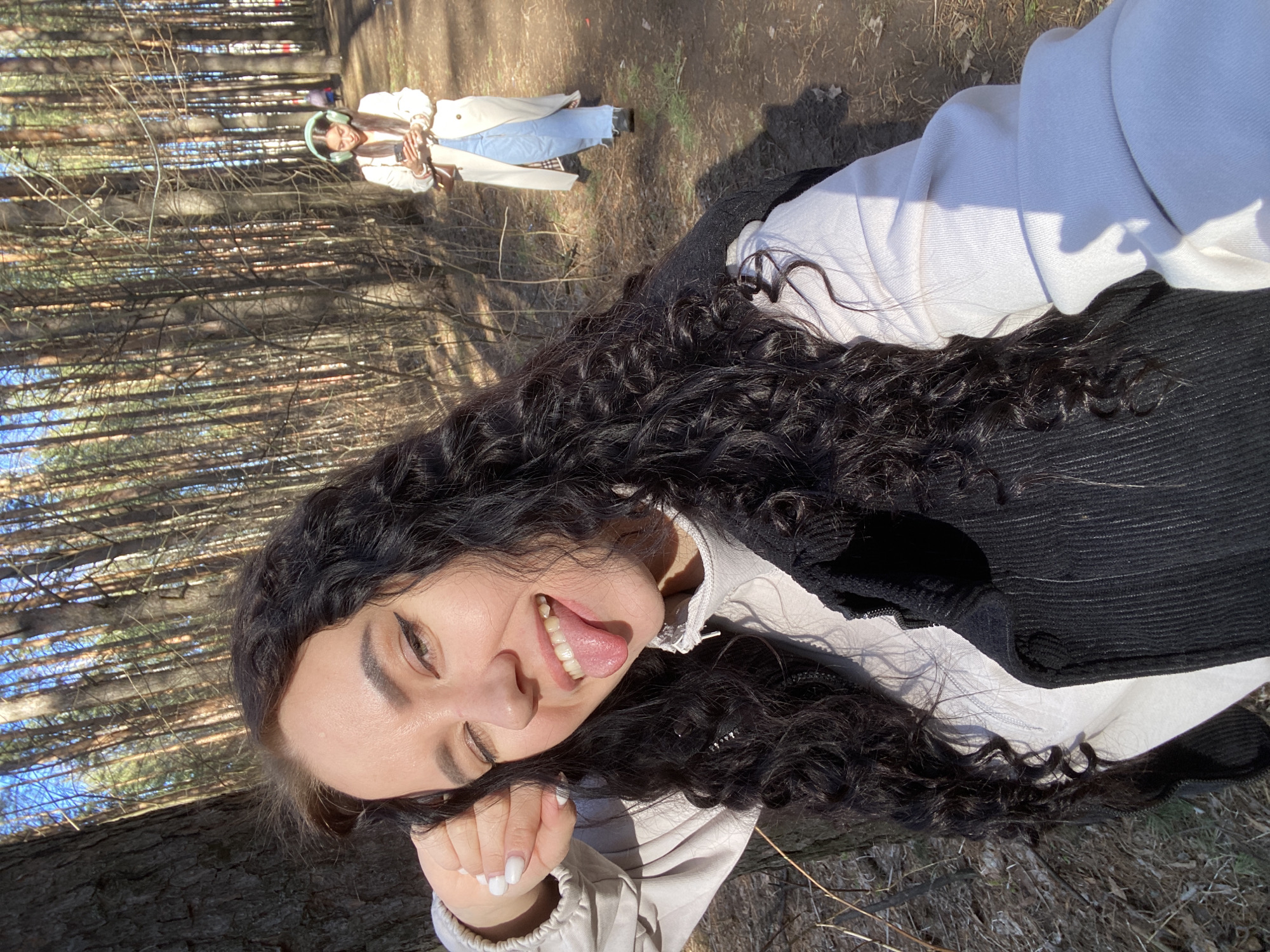 The image depicts two women in a forest setting, potentially located in or near the city of Chaikovsky, Russia. The primary focus is on the woman in the foreground, who is positioned in a recumbent pose. She has long, dark, curly hair and is wearing a white top with a black vest or jacket. She is smiling, with her tongue sticking out. Her left hand, featuring a white manicure, is supporting her head.
In the background, a second woman is visible standing among the trees. She is wearing light-colored jeans, a white coat, and light blue headphones. She appears to be holding a phone.
The environment is a wooded area with numerous trees, with light shining through the canopy creating shadows on the forest floor. The ground appears to be covered with leaf litter and other natural debris.