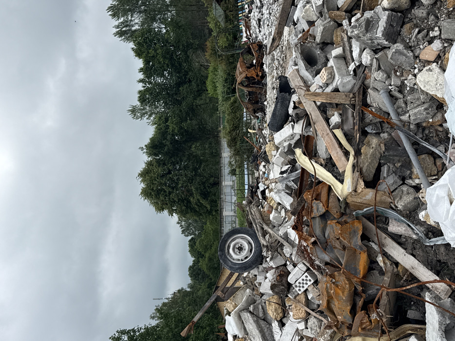 Debris and rubble dominate the foreground, consisting of broken concrete, bricks, and rusted metal. A tire lies amidst the rubble, suggesting a vehicle was involved in the destruction. A few charred remains indicate fire damage. In the background, a line of green trees separates the debris from a gray, cloudy sky. The scene evokes a sense of devastation and destruction.