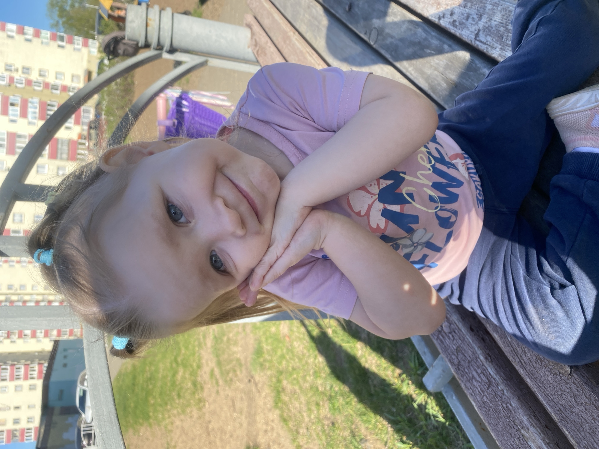 A young girl with blonde hair, styled with small blue hair ties, lies on a wooden bench in what appears to be a park in Chaikovsky, Russia. She is wearing a pink t-shirt with a graphic design and dark blue pants. Her hands are positioned supporting her face, and she is smiling. In the background, a multi-story apartment building with a light colored facade and red window accents is visible. Patches of green grass are present in the foreground and background. The bench is situated near a playground structure with metal arches. The lighting suggests it is daytime, with shadows cast on the bench and ground.