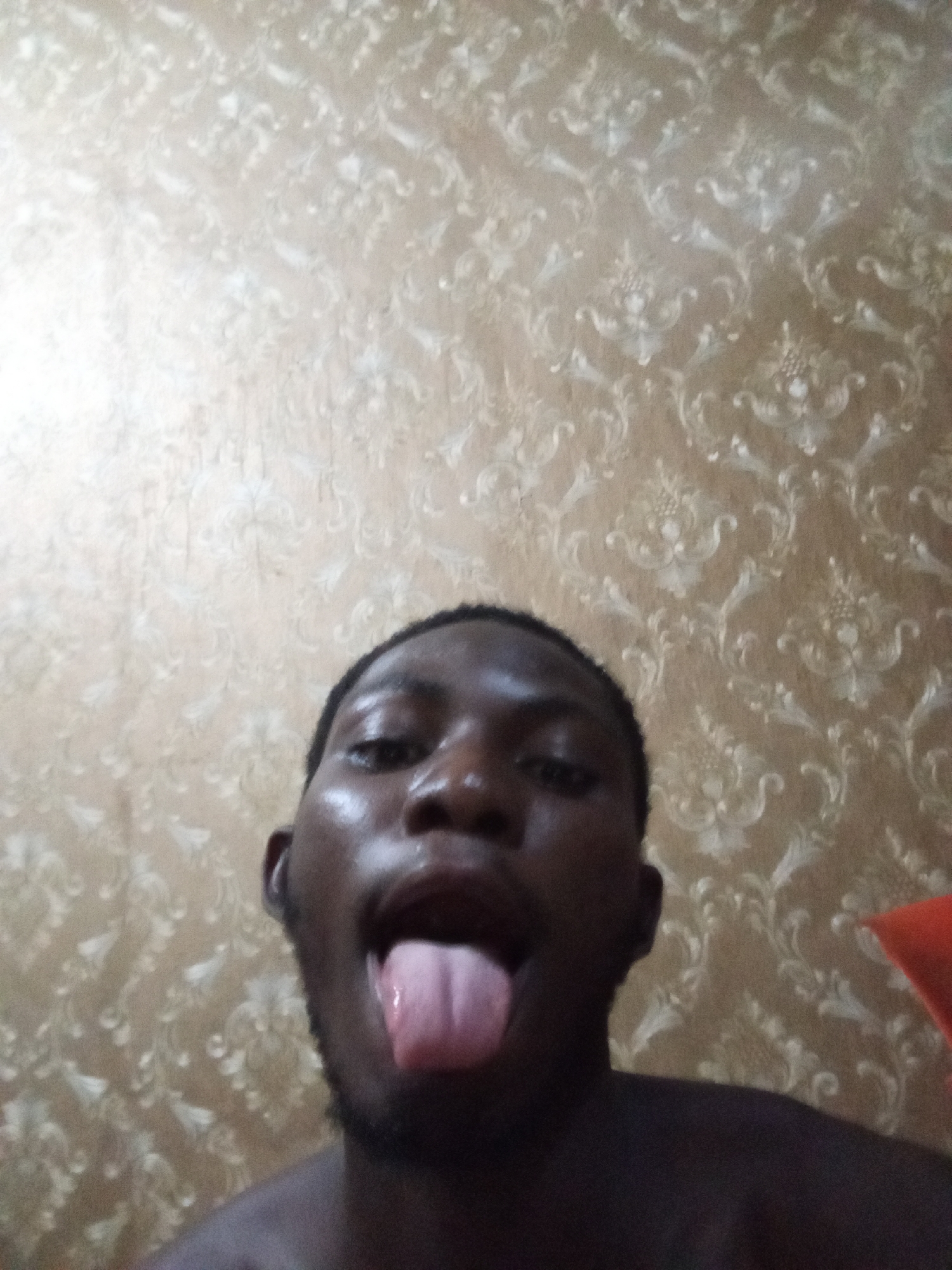A dark-skinned man is sticking his tongue out at the camera. He has short black hair and a beard. The background is a patterned wallpaper. The location is Unknown, Unknown.