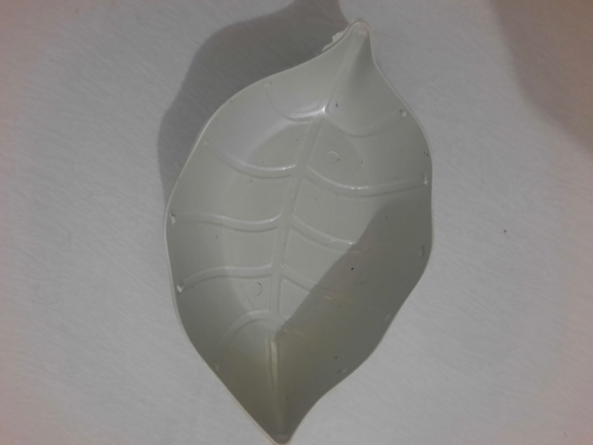 A white, leaf-shaped plastic dish sits on a textured white surface, possibly a tablecloth or a wall. The dish has embossed veins and a slightly curved shape, designed to hold small items. The lighting casts a shadow to the left of the dish. The overall scene is simple and domestic, with no discernible connection to Lhokseumawe, Indonesia.