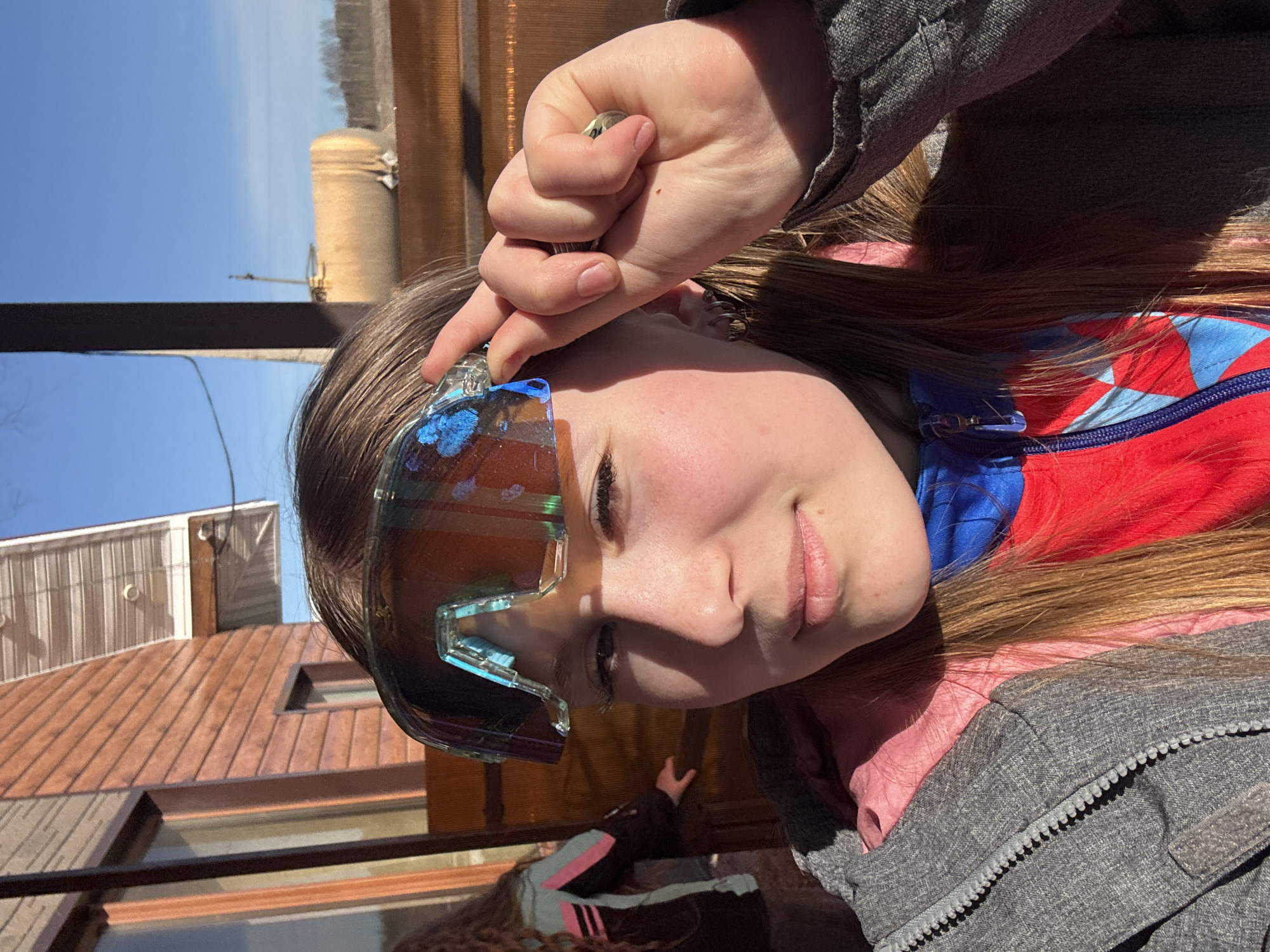 A young person with light skin and light brown hair is posing for a selfie. They are wearing futuristic-looking sunglasses with blue accents. Their left hand is raised near their forehead, partially obscuring the top of the glasses. They are wearing a gray jacket over a pink shirt and a red and blue zip-up top.
The environment outside features architectural structures, including a building with a corrugated metal roof. Visible in the background is a cylindrical tank, likely part of an industrial complex. The sky is clear and blue. The specific location within Gory, Russia, cannot be determined based solely on this image.