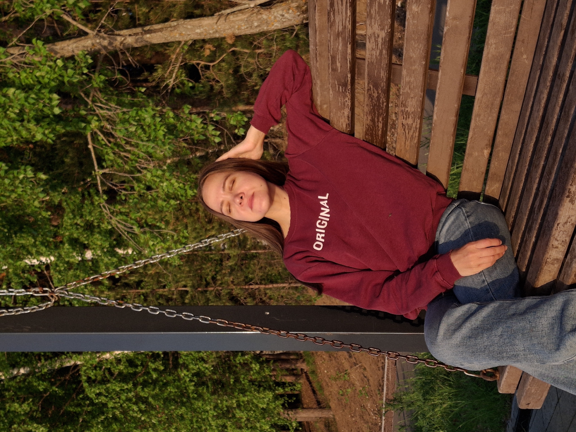 A young woman is reclining on a wooden bench in an outdoor setting. She is wearing a maroon sweatshirt with the word "ORIGINAL" printed vertically on the chest, and blue jeans. She is resting her head on her hand.
The bench appears to be a swing bench, suspended by chains visible in the image. The backdrop consists of trees and foliage, suggesting a park-like environment.
The location is Chaikovsky, Russia.
