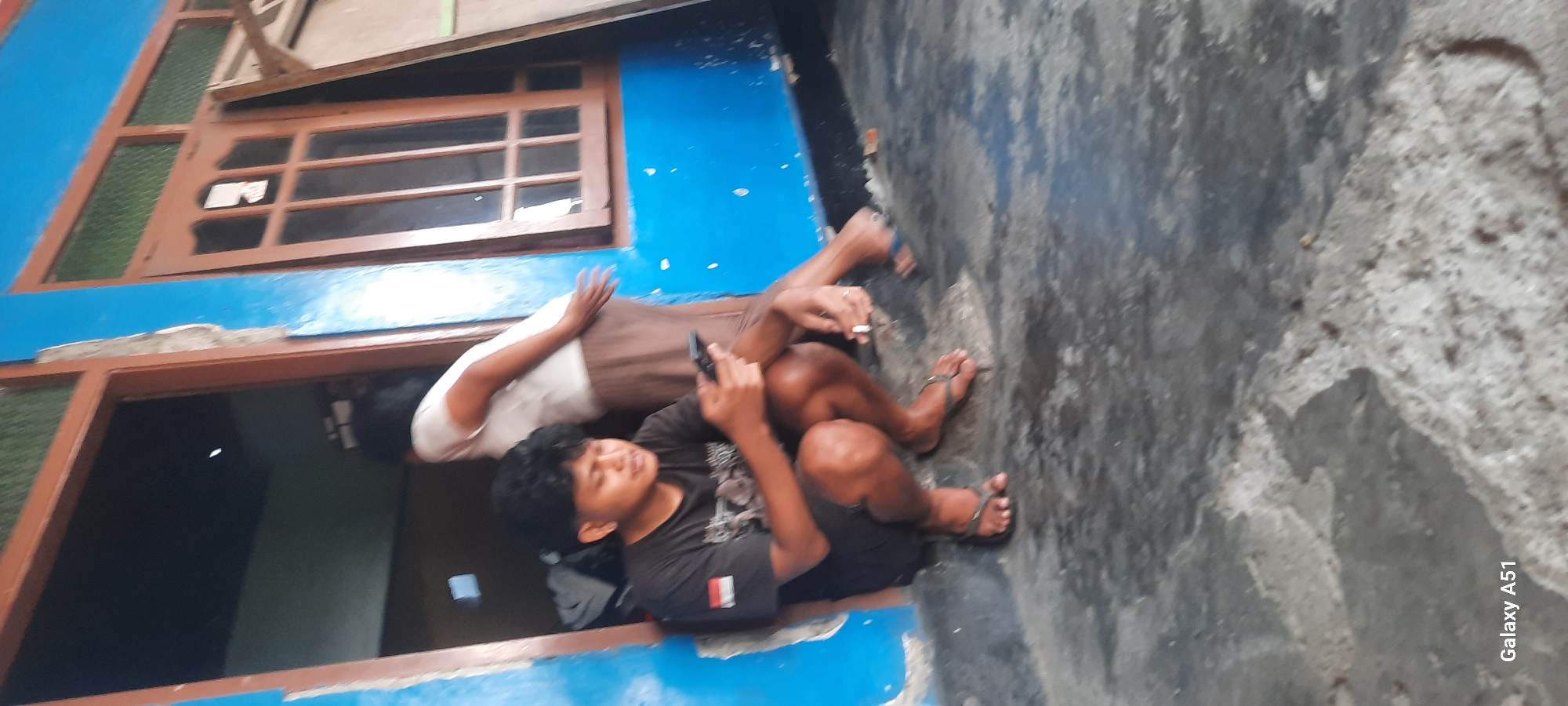 A young man is crouching on a concrete porch in front of a house. He is wearing a black t-shirt with a graphic print and black shorts, along with flip-flops. He is holding a mobile phone in his right hand and appears to be interacting with it. A cigarette is visible between the fingers of his left hand. Behind him, another person, partially visible, stands in a doorway wearing a white t-shirt and brown pants. The house has a blue painted exterior with wooden window frames. The ground is made of rough concrete, and there are some water stains. The image appears to be taken outdoors in a residential setting, possibly in Selopuro, Indonesia, given the "Galaxy A51" watermark, a phone model commonly used in the region.