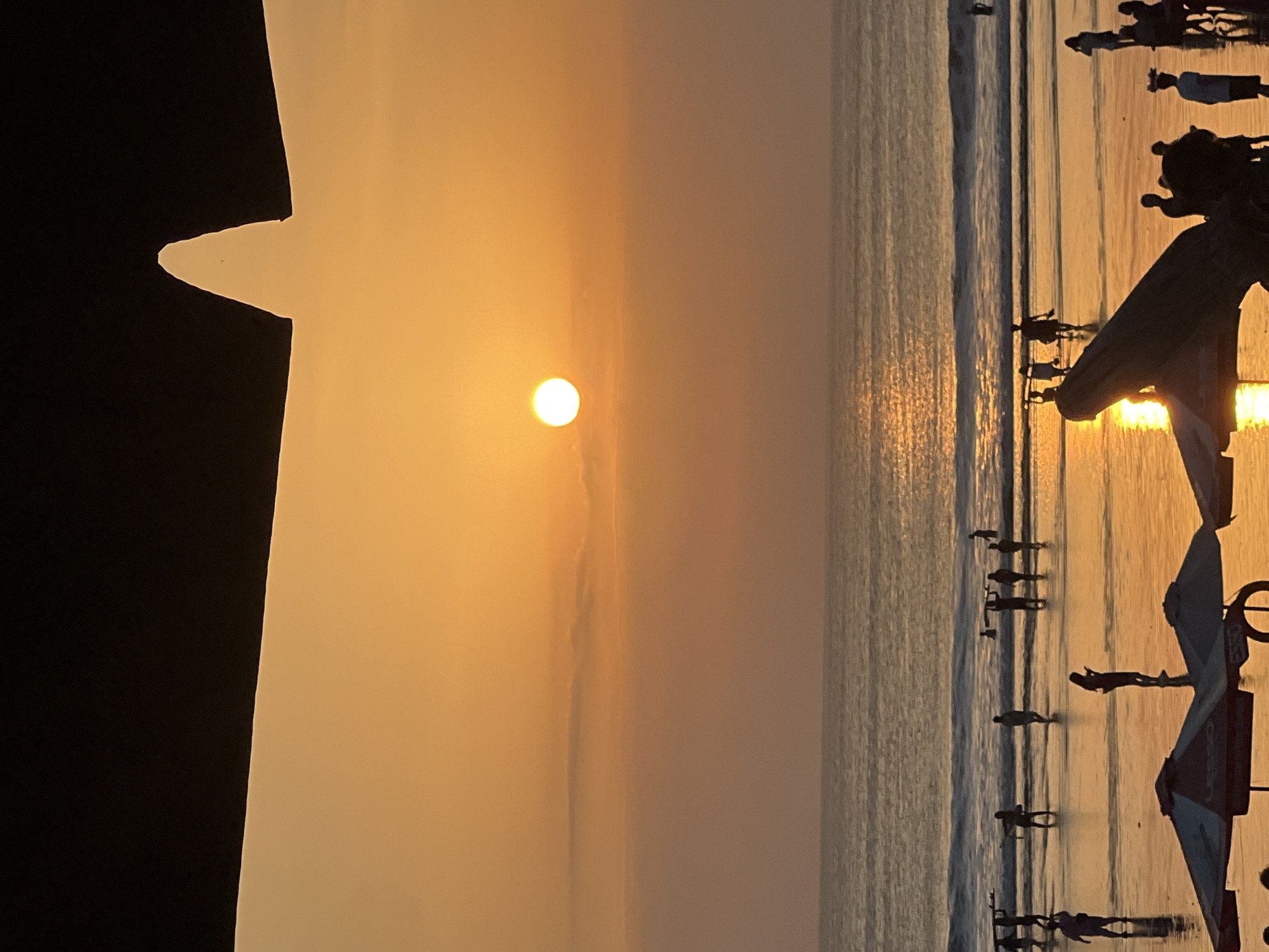 This image captures a vibrant sunset scene at Kuta Beach in Kuta, Indonesia. The sky is a warm gradient of orange and yellow, with the sun prominently positioned in the center, casting a shimmering reflection on the ocean. Silhouettes of people are visible on the beach, some carrying surfboards, suggesting activities like surfing or simply enjoying the sunset. The presence of beach umbrellas and a large piece of driftwood in the foreground indicates a typical beach setting. The overall atmosphere appears to be one of leisure and natural beauty, characteristic of a popular tourist destination like Kuta.