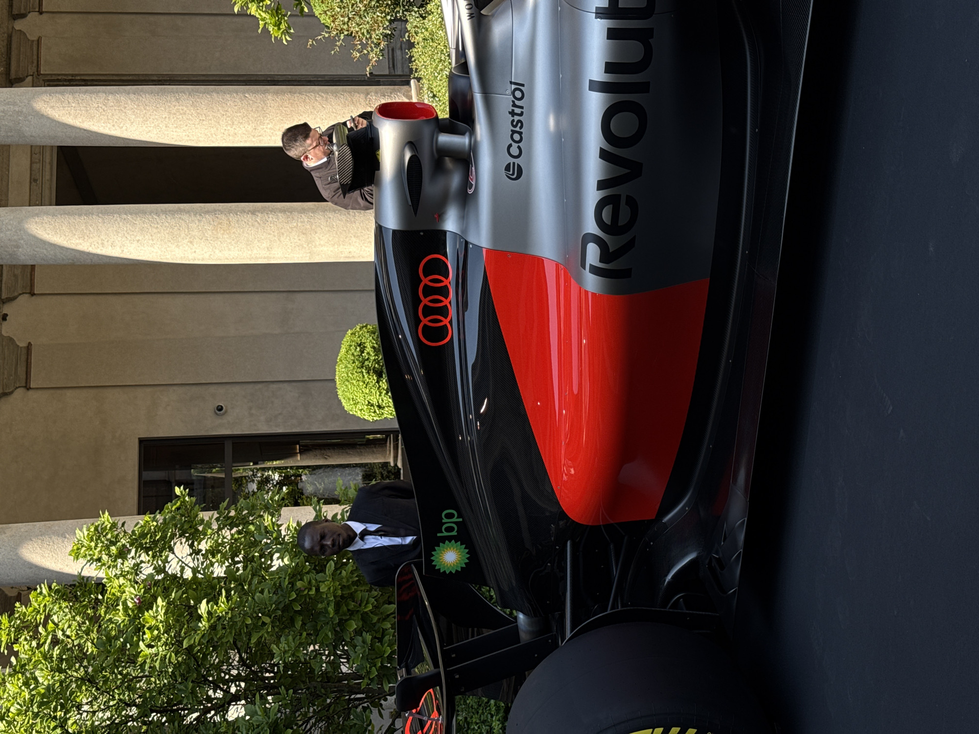 An outdoor shot captures a sleek, modern race car on display in what appears to be a European city courtyard, consistent with Milan, Italy. The car features a distinctive black, red, and silver livery, adorned with prominent sponsor logos including the red "Audi" rings on the nose, "Castrol" and "Revolut" along the sidepod, and the "bp" green sunburst logo near the rear wheel. Parts of the car, like the side panels and rear wing, show exposed carbon fiber.
Two men are visible in the scene. Near the front of the car, an older man with glasses, wearing a dark jacket, is intently focused on the vehicle, possibly holding a small electronic device like a phone or camera. Further back, partially obscured by green foliage and the car's rear wing, a younger Black man in a dark suit and white shirt stands, looking towards the car.
The background reveals classical architecture with large, light-colored stone columns casting long shadows, suggesting bright daylight. Behind the columns, part of a building facade with windows and what looks like a security camera can be seen. Lush green bushes and trees frame the scene, with some foliage partially obscuring the man in the suit. The bright sunlight indicates a clear day, likely late morning or early afternoon. The stationary car and the observers suggest an exhibition, launch event, or public display.