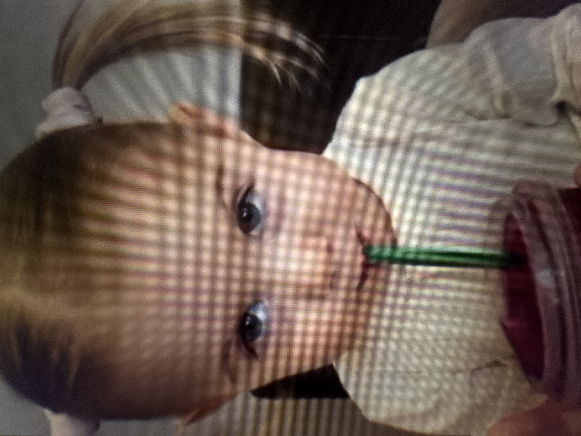 A rotated medium close-up image shows a baby or toddler drinking from a purple cup with a green straw. The child, positioned on their side with their head to the left and body to the right of the frame, looks directly at the camera with blue eyes and a calm, slightly smiling expression. Their light brown hair is pulled into a high ponytail on top of their head, secured by a light-colored scrunchie. They are wearing a light-colored, possibly white or cream, ribbed top. The purple cup, held by the child, contains a dark red liquid, and the bright green straw is visible entering their mouth. The background is dark and out of focus, suggesting an indoor setting. The overall image displays a visible pixel grid, indicating it is likely a photograph taken of a digital screen. No text is visible in the image.