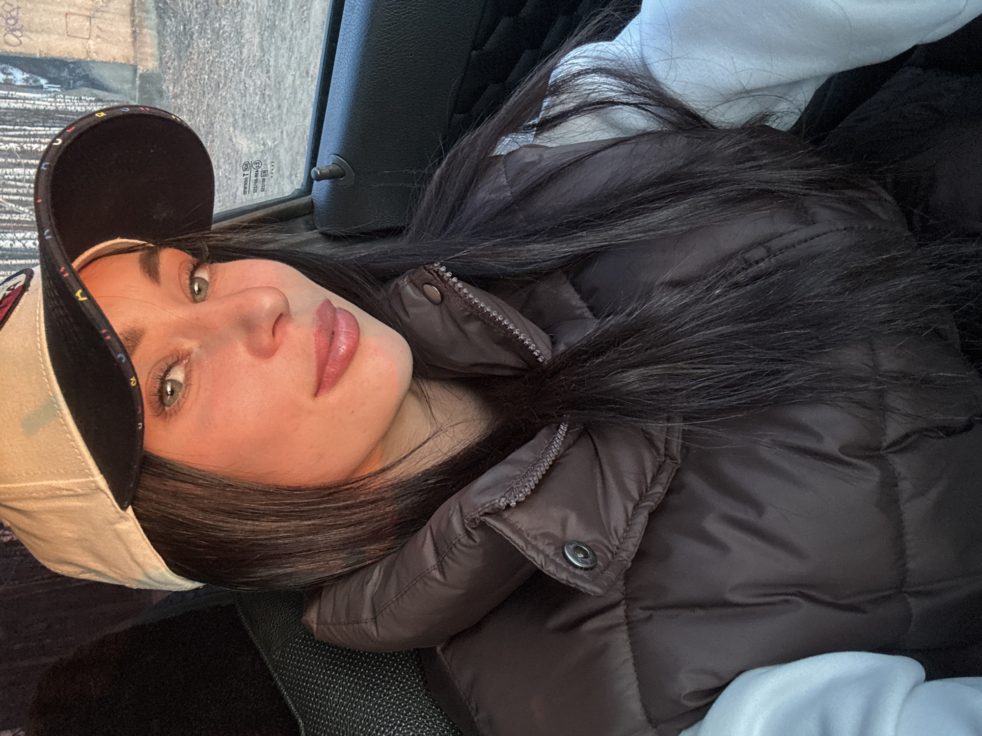 The image depicts a young woman wearing a black and white baseball cap, and a dark vest, seated in what appears to be a vehicle. Her hair is dark, and she is looking towards the camera. The background includes a car window and the interior of the vehicle. The location is not discernible as Chaikovsky, Russia or any other specific location.