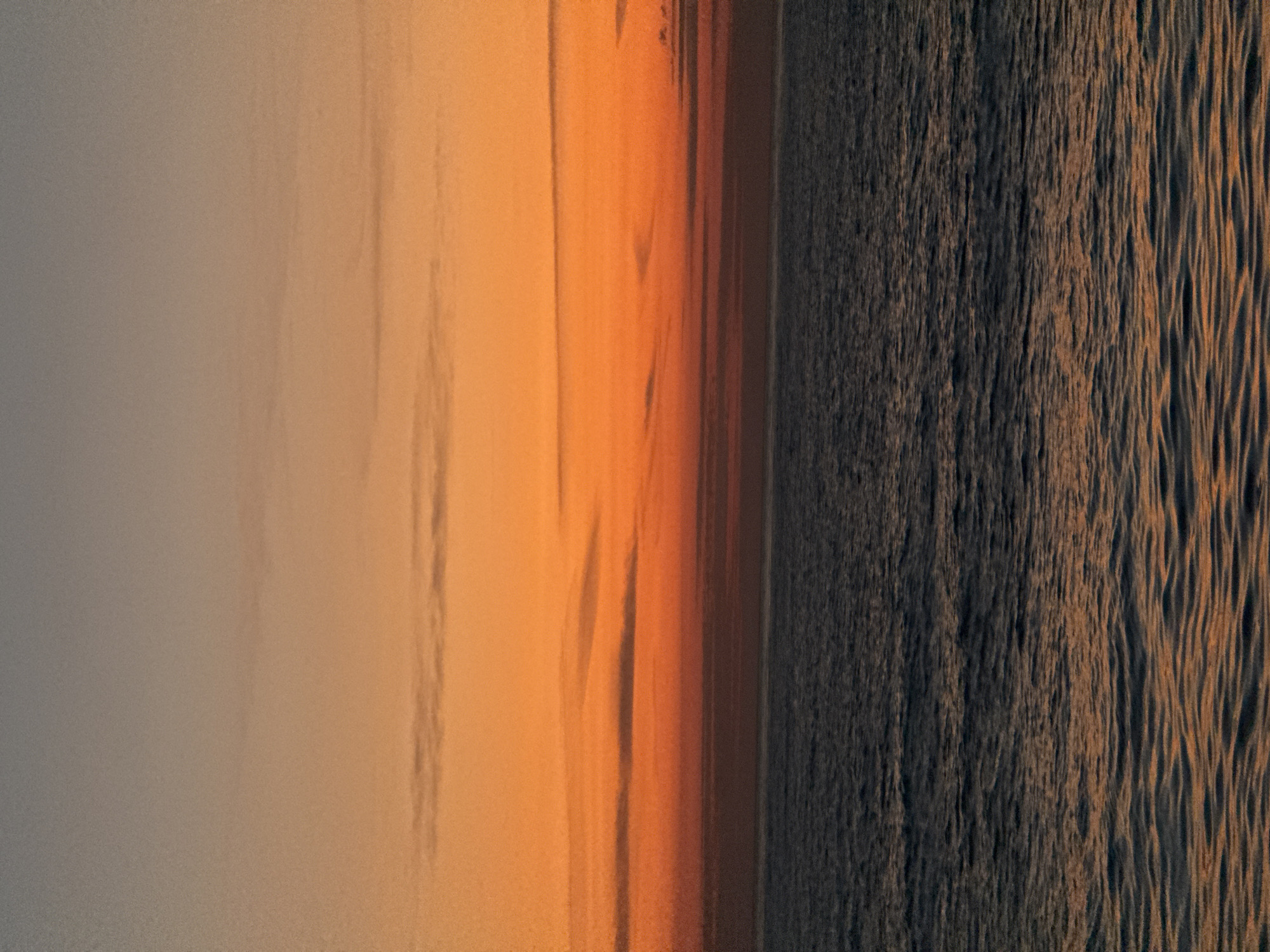 The image depicts a body of water, likely the Black Sea, during sunset. The sky transitions from a muted gray on the left to shades of orange and brown towards the center, suggesting the setting sun. The sea displays dark, textured ripples catching the fading light. The scene location cannot be determined based solely on the image content. Therefore, the location in the city of Batumi, Georgia, is not evident.