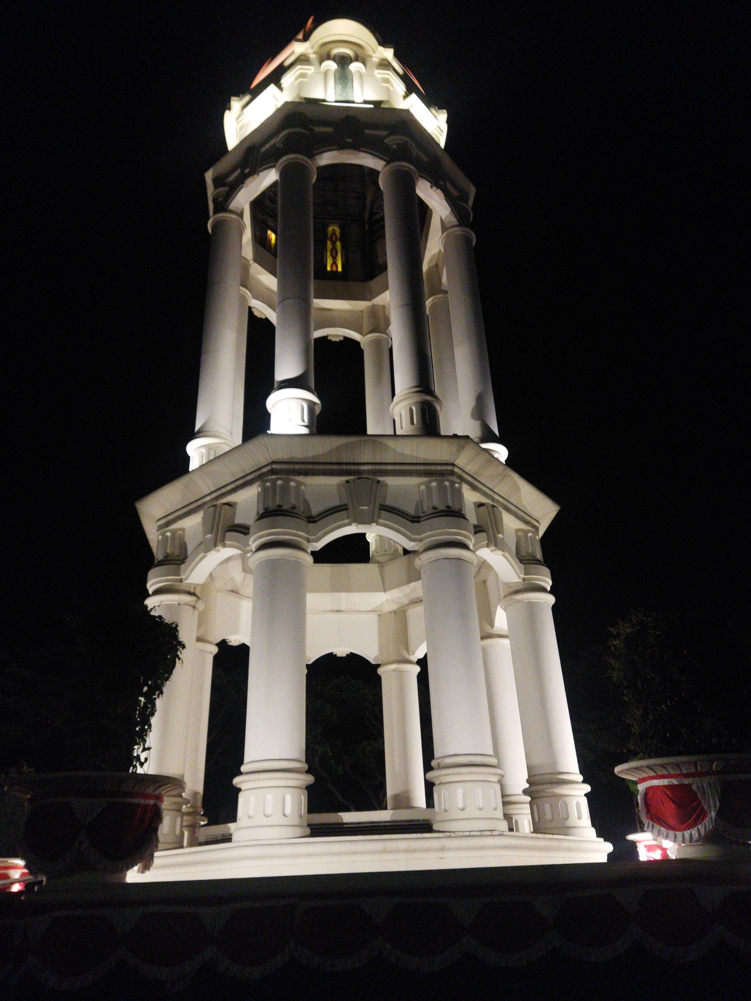 This is a low-angle, nighttime photograph of the Tugu Muda monument in Semarang, Indonesia. The white concrete monument, illuminated by spotlights, stands tall against a dark sky. It features a central cylindrical structure surrounded by several large, fluted columns. The monument has multiple levels with arched openings and decorative architectural elements. In the foreground, there are two large, round planters adorned with red and white fabric. The ground in front of the planters is covered with a dark fabric or banner. Trees are visible on either side of the monument. The lighting creates strong shadows and highlights, emphasizing the monument's architecture.
