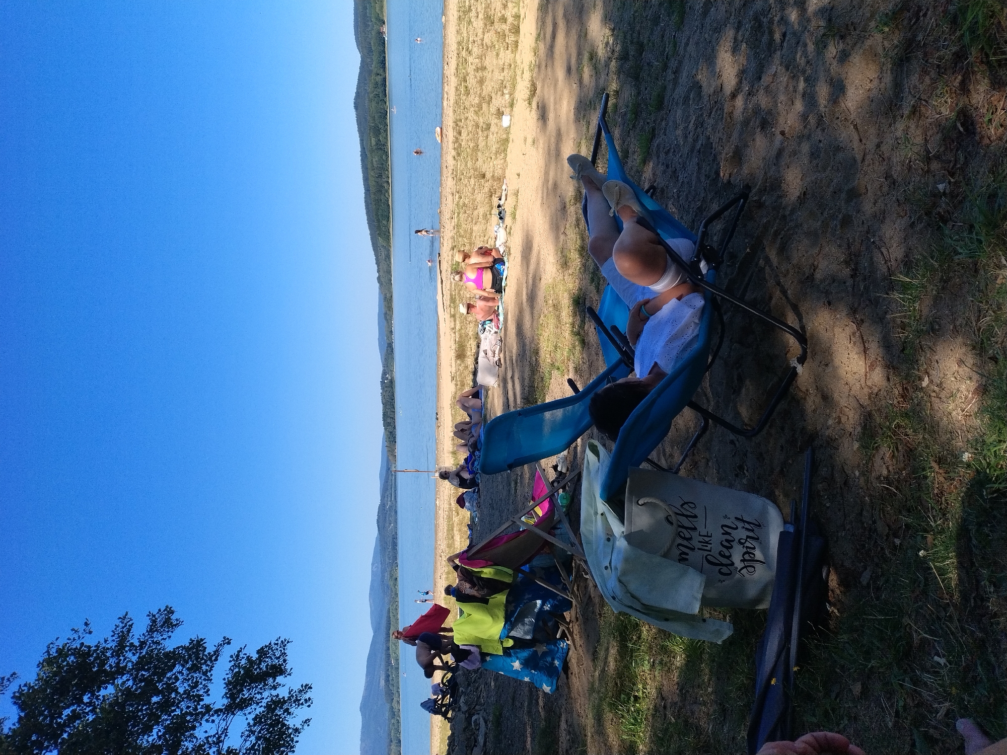 The image depicts a sunny day at a beach or lakefront in Radslav, Czechia. In the foreground, a person is relaxing in a blue reclining chair, wearing white shorts and a white top. Another blue reclining chair is positioned nearby. A canvas tote bag with the inscription "smells like clean spirit" rests on the ground next to the bag. Further back, several other people are visible on the sandy shore. Some are seated or lying down, while others are near the water's edge, with a few individuals swimming or wading. The lake stretches out towards a tree-lined horizon, with hills in the distance under a clear blue sky. The overall scene is one of leisure and recreation on a warm day.
