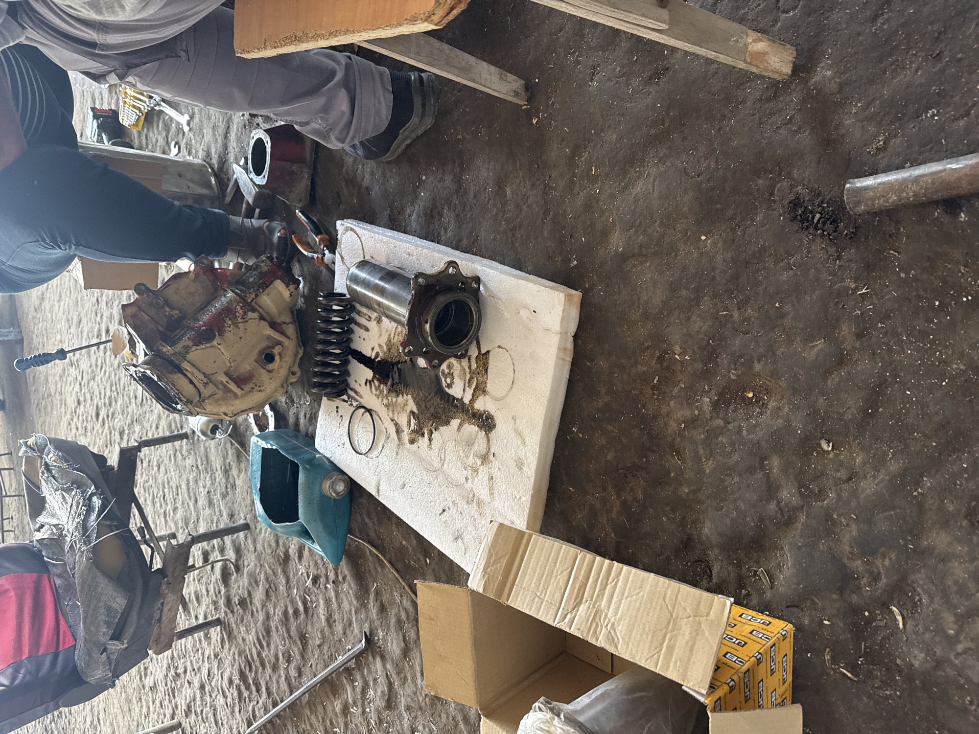 The image depicts an indoor scene within what appears to be a workshop or garage in Sarnevo, Bulgaria, where mechanical work is in progress. Two individuals are partially visible: one wearing light gray pants and a matching top, and another in dark pants and a dark long-sleeved shirt, with their hands near the components.
The dirty, earthy floor is covered with various tools and machine parts. A large, greasy, and somewhat rusty mechanical component, possibly a differential or gearbox housing with remnants of red paint, lies prominently. Next to it, a metallic cylinder with a flanged end and a large coil spring are resting on a white polystyrene foam block, which is stained with oil and grease. Smaller metal rings and other tools, including pliers and a screwdriver-like implement, are scattered nearby. A cut-open, light blue plastic container is also on the floor.
In the background, wooden planks and beams are visible. An open cardboard box and another smaller cardboard box, clearly displaying the text "JCB" multiple times, are present. The scene suggests the repair or assembly of heavy machinery components in a functional and utilitarian environment. The lighting is consistent with an indoor setting.
