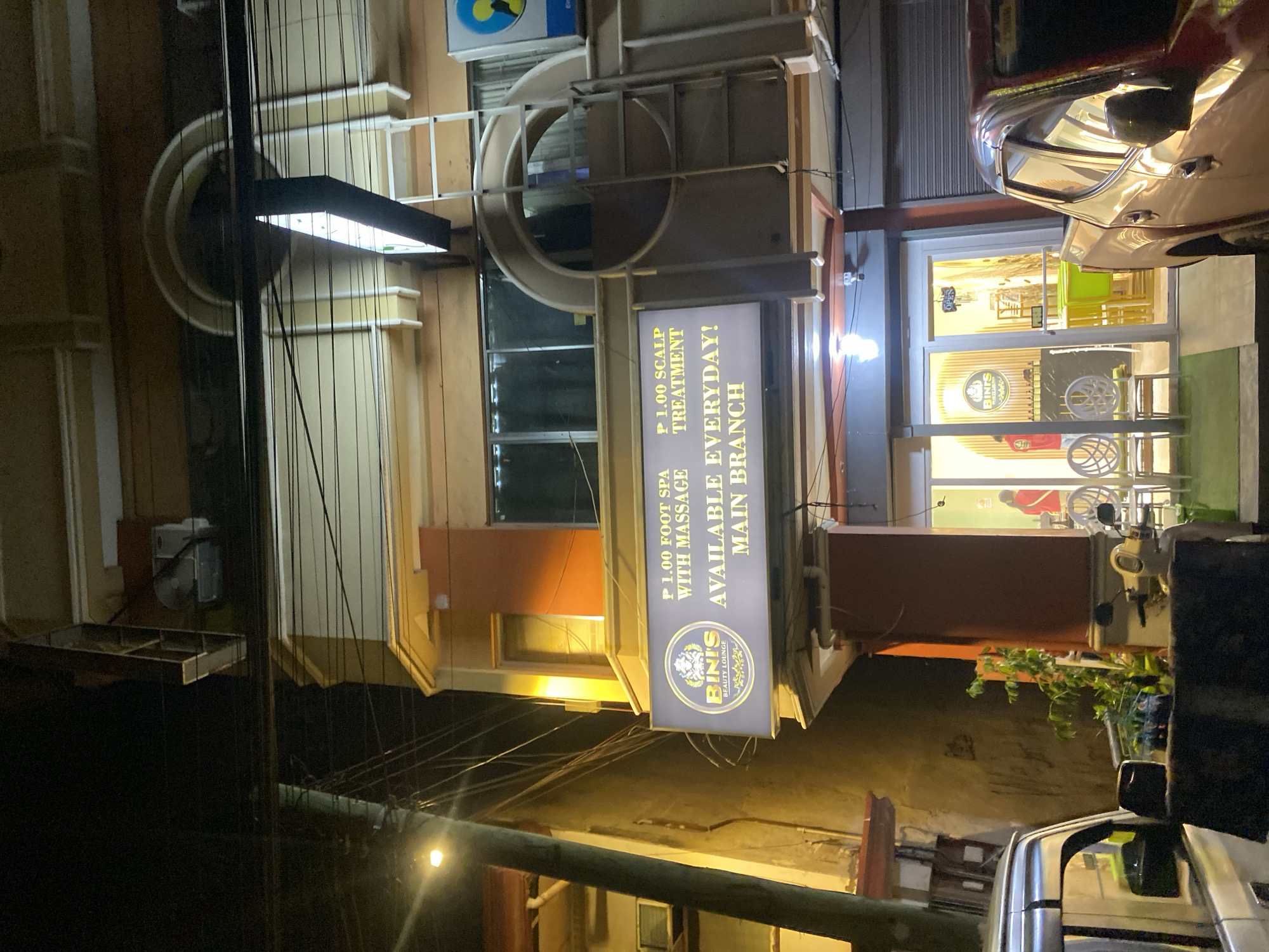 A nighttime, eye-level exterior shot captures the front of a commercial establishment in Davao City, Philippines. The building is a multi-story structure with an orange and cream facade. A prominent sign, illuminated with yellow lettering, displays "BIN'S BEAUTY LOUNGE" and advertises "P 1.00 FOOT SPA WITH MASSAGE" and "P 1.00 SCALP TREATMENT AVAILABLE EVERYDAY! MAIN BRANCH". The entrance to the establishment is a glass door with "OPEN" visible inside. To the left of the entrance, two white, stylized chairs are placed on the sidewalk. A silver pickup truck is parked in the foreground on the left, and a red pickup truck is visible on the right. A white scooter is parked near the entrance. Power lines are visible across the top of the frame. The overall scene depicts a commercial street in Davao City at night.