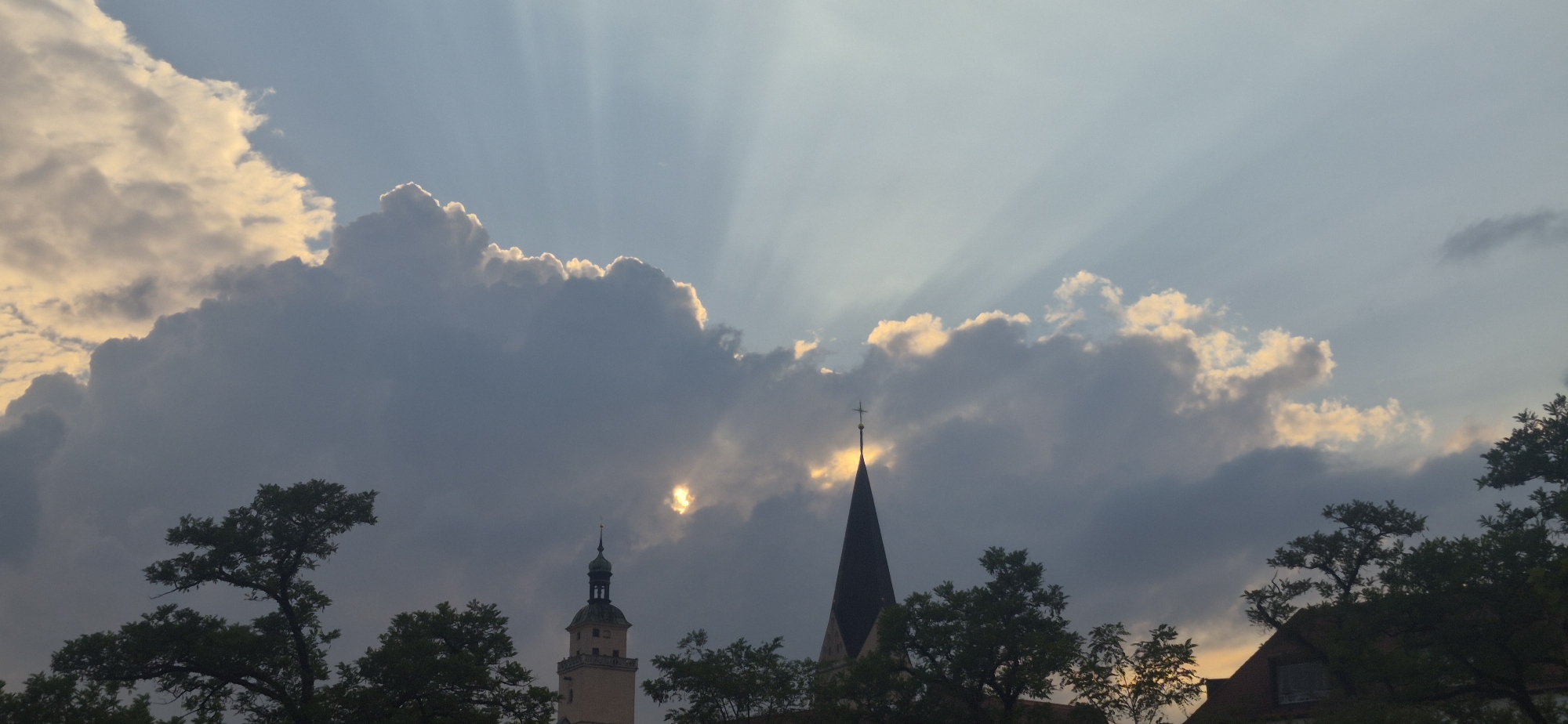 This image depicts a dramatic sky with sunbeams breaking through dark clouds. In the foreground, silhouetted trees and the spires of two church towers are visible. The church on the left has a lighter, square tower topped with a green, pointed roof. The church on the right features a dark, slender, pointed spire. The sun is partially obscured by a cloud, creating a bright, glowing effect. The scene suggests a late afternoon or early evening setting. Based on the architecture of the church towers, particularly the one on the left which resembles the "Neues Schloss" tower in Ingolstadt, Germany, this scene is likely located in the city of Ingolstadt. There are no people or animals visible in the image.