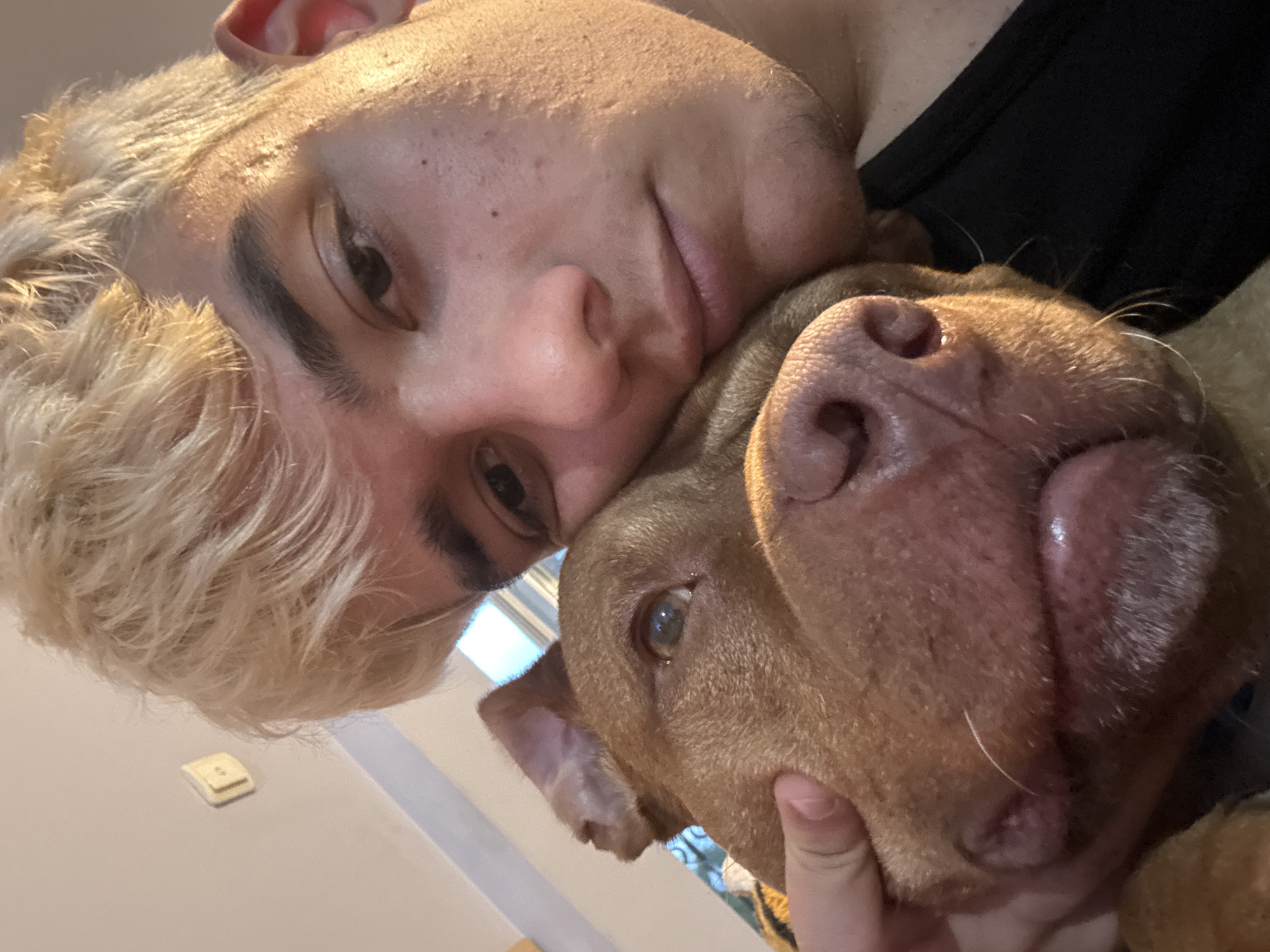 An indoor selfie captures a person with dyed blonde hair embracing a dog with brown fur. The person's left hand is visible, gently touching the dog's head. The background shows a plain wall with a small light switch. The lighting suggests a warm, possibly evening, atmosphere. The location is not specified in the image or its metadata.