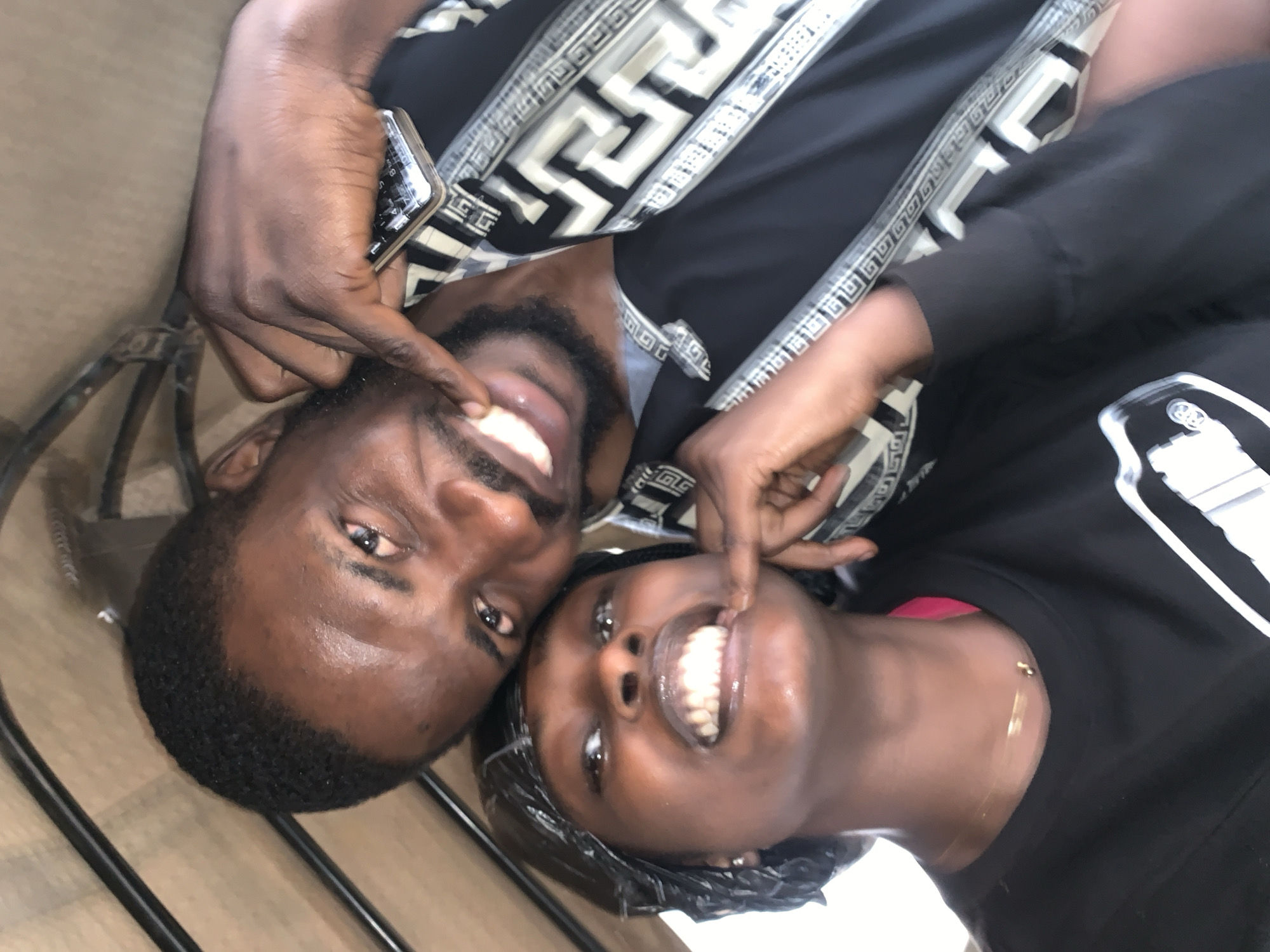 The image captures a close-up, selfie-style shot of two individuals, a man and a woman, likely in a vehicle. The man, with dark skin and short, dark hair, is in the foreground, leaning in towards the camera. He has a beard and is smiling widely, with his right index finger touching the corner of his mouth. He is wearing a black shirt with a distinctive white and black geometric pattern along the collar and sleeves. A silver-colored mobile phone is visible in his right hand, resting on his chest.
The woman, also with dark skin and dark hair covered by a black headscarf or bonnet, is positioned slightly behind and to the left of the man. She is also smiling broadly, revealing her teeth. Her left index finger is touching the corner of her mouth. She is wearing a black long-sleeved shirt. A delicate gold necklace is visible around her neck.
The background appears to be the interior of a vehicle, possibly a tricycle or a car, with metal bars and beige-colored lining visible. The lighting suggests an outdoor setting during the day. The image has a tilted perspective, common in selfies. The overall mood is playful and affectionate. The image is not specific to any particular location within the city of Jos, Nigeria, and provides no distinct landmarks or contextual clues that would allow for such a determination.