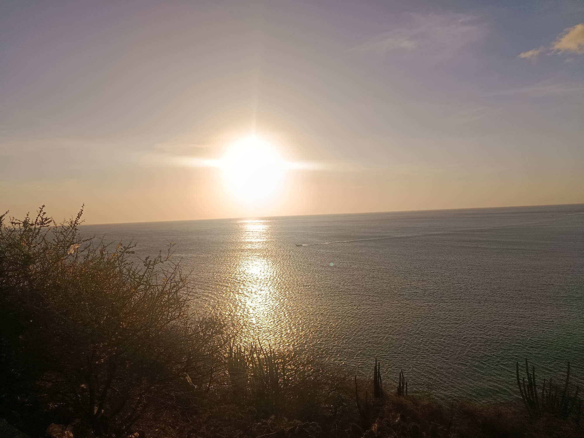 The image captures a coastal scene at sunset in Juan Griego, Venezuela. The sun is positioned centrally, reflecting intensely on the ocean surface, creating a shimmering path of light. A small boat is visible on the water. The foreground shows land with dry vegetation, including shrubs and cacti. The sky transitions from a pale orange near the horizon to a light blue at the top.