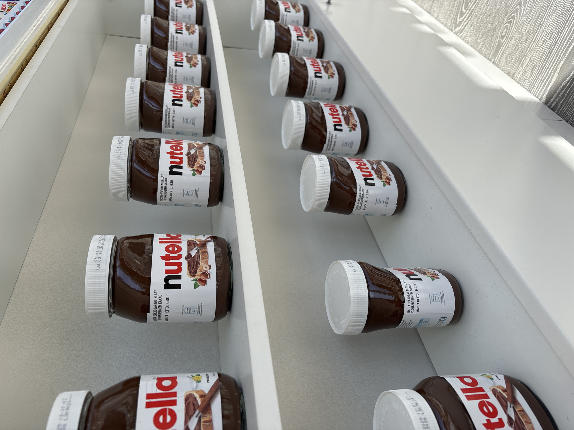 The image shows rows of Nutella jars on a white shelf. The jars have white lids and labels with the Nutella logo and product information in Cyrillic script. The label identifies the product as hazelnut paste with cocoa, net weight 630g. A date code "LU 02-11 2025 13:01" is printed on the jar lids. The shelf appears to be part of a retail display. The top left corner of the image shows the label of a "Kinder" product. Based on the product information in Cyrillic, the scene is likely in Russia, and due to the reference to "Sochi" in the request, it is inferred that the scene is in the city of Sochi, Russia.