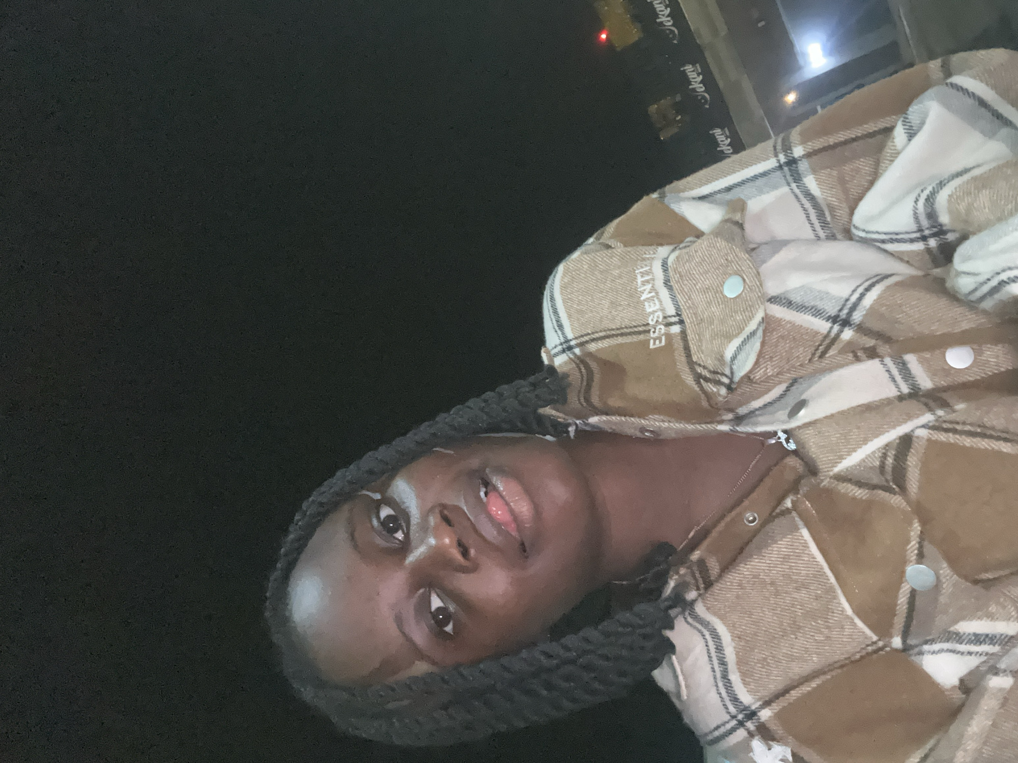 The image presents a close-up, chest-up view of a dark-skinned individual, likely female, posing for a photo, presumably a selfie, in an outdoor nighttime environment. The subject has dark, braided hair and is looking directly at the camera with a subtle expression, lips slightly parted, revealing the tip of her tongue. Her skin shows a reflective sheen on the forehead, nose, and cheeks, possibly from ambient lighting. She is wearing a collared button-up shirt featuring a plaid pattern in tones of brown, tan, and off-white. The shirt has silver-colored snap buttons and a pocket on the upper left side (from the viewer's perspective, the subject's right), which bears the embroidered word "ESSENTI" in white, suggesting a brand name such as "ESSENTIALS." A thin silver-colored chain necklace with a small pendant is visible around her neck.
The background is largely obscured by darkness, indicative of night. To the right of the subject, several illuminated signs are discernible. The most prominent displays the word "Jekani" multiple times in white, stylized script against a dark panel. Below this, additional white lettering is partially visible, likely part of the same establishment's signage. Further in the distance, a bright, rectangular white light source and a small red light are visible. The presence of the "Jekani" sign strongly indicates the location is in or around Owerri, Imo State, Nigeria, which includes the town of Ihiagwa, where Jekani Hotels and Suites is known to operate.