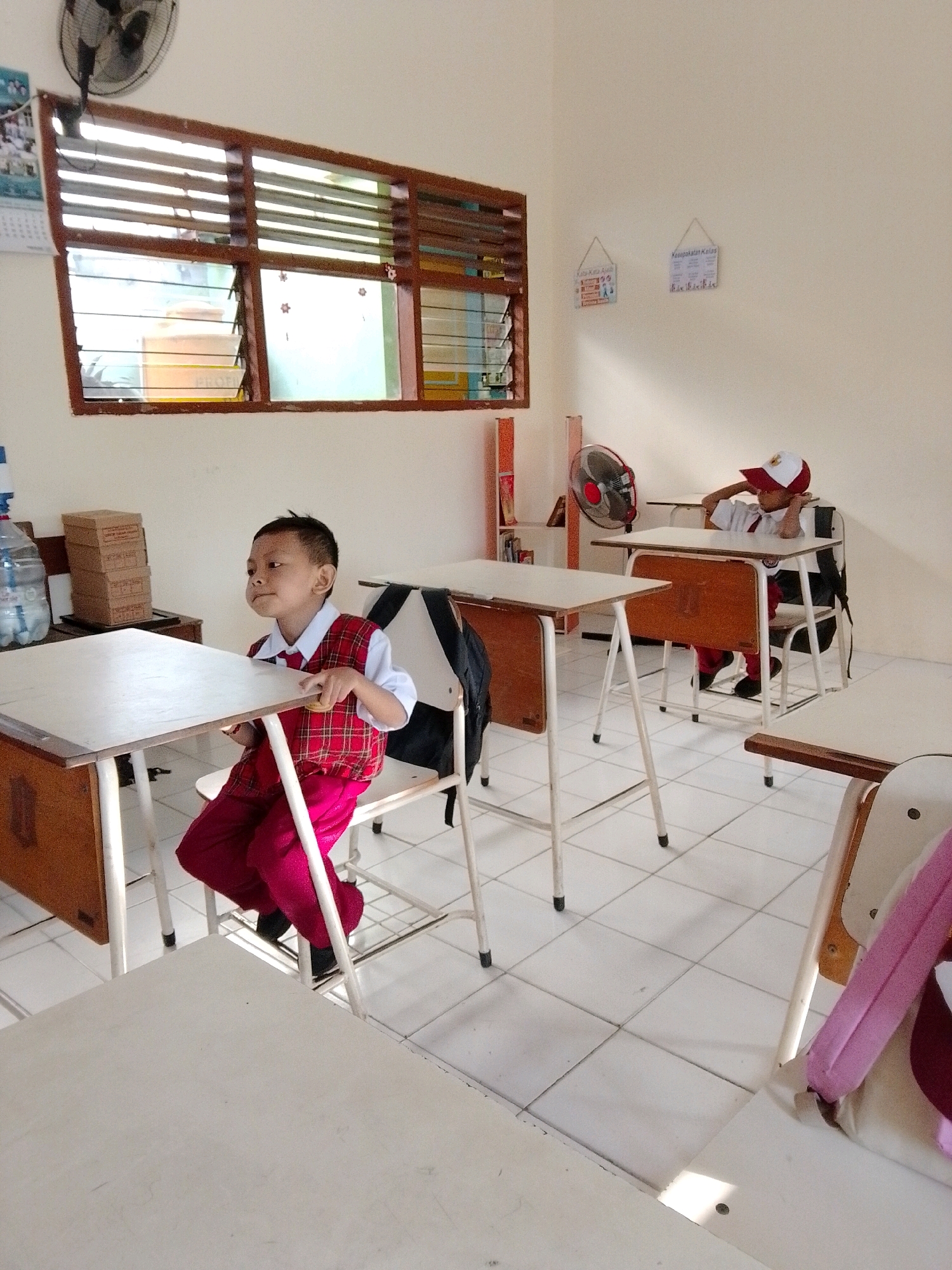 The image depicts a classroom setting. In the foreground, a young boy wearing a red and white checkered vest over a white shirt and red pants sits at a desk. He appears to be looking to his left with a thoughtful or perhaps bored expression. His hands are resting on the edge of the desk. Behind him, another student, wearing a white shirt, red pants, and a red and white cap, is seated at a desk with their head resting on their hand, appearing to be dozing or deep in thought. The classroom has white tiled floors and cream-colored walls. There are multiple wooden desks with metal frames, chairs, and a few personal items like a backpack and a water bottle. A window with wooden blinds is visible on the left wall, and two framed posters hang on the right wall. A ceiling fan is mounted on the wall in the upper left corner. The overall impression is that of a typical, somewhat sparsely decorated elementary school classroom. Without further context, it is impossible to definitively place this scene in Surabaya, Indonesia. However, the uniforms worn by the children, the style of the classroom, and the script on the posters (which appear to be in Indonesian) are consistent with educational institutions in Indonesia.
