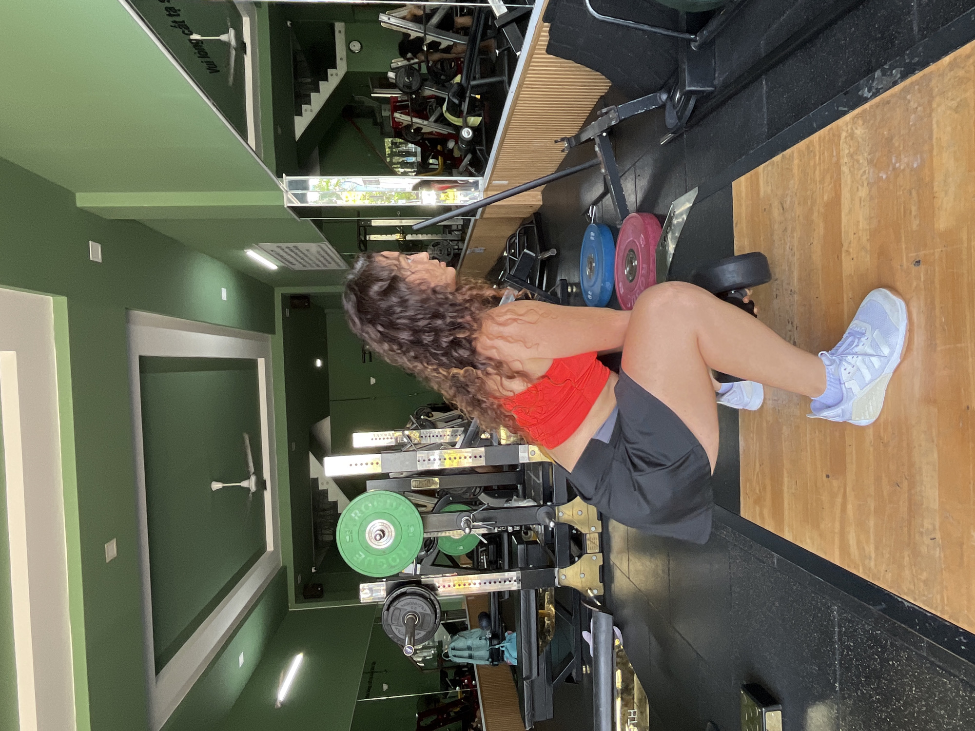 In a gym in Phan Thiết, Vietnam, a woman with curly brown hair is performing a squat. She is wearing a red sports bra and black shorts, with light purple sneakers. She is holding a barbell with weights on each end, and her body is positioned in a squatting stance. The gym is equipped with various weightlifting machines, weight plates, and mirrors. The walls are painted green, and the ceiling has a unique design with green and white accents. A ceiling fan is visible in the upper portion of the image. The floor consists of black rubber mats and a wooden platform where the woman is exercising. Reflections in the mirrors show other gym equipment and people working out.
