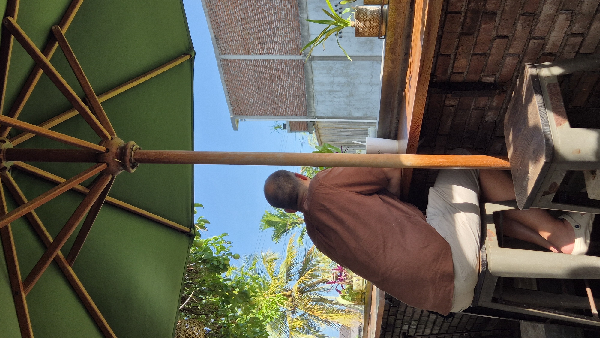 A photograph taken in Pekutatan, Indonesia, shows a person sitting at a wooden table under a green umbrella. The person has a shaved head and is wearing a brown shirt and light-colored shorts. The table is next to a brick wall. In the background, there are palm trees and buildings. The sky is blue.