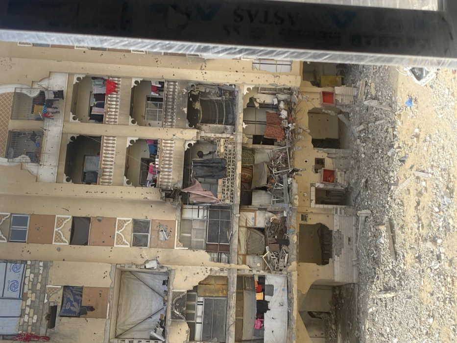 The image displays a severely damaged multi-story residential building in Khan Yunis, Palestinian Territory. The building's light-colored facade exhibits extensive destruction, characterized by numerous small, circular impact holes indicative of projectile strikes across all visible floors. Large sections of the external walls, particularly on the lower and central levels, are either completely missing or heavily compromised, exposing the ruined interiors of multiple apartments. Windows are shattered or entirely absent.
Despite the widespread damage, evidence of habitation persists: multiple balconies on the upper-middle floors have laundry hanging to dry, including red, pink, and multi-colored garments. Some open spaces and missing window areas are covered with improvised materials, such as tarps and sheets; a blue tarp on the third visible floor from the bottom, center-left, displays a faint UNICEF logo. Within the exposed rooms, debris, fragmented furniture, and remnants of household items are visible.
The ground directly in front of the building is covered by a dense field of rubble, composed of broken concrete, twisted rebar, and other construction materials. Two distinct red, cube-like structures are visible at ground level, flanking a heavily damaged central area. A partial view of a metallic vertical structure, bearing the text "ASTAS," frames the extreme right foreground of the image. No individuals are visible in the scene.