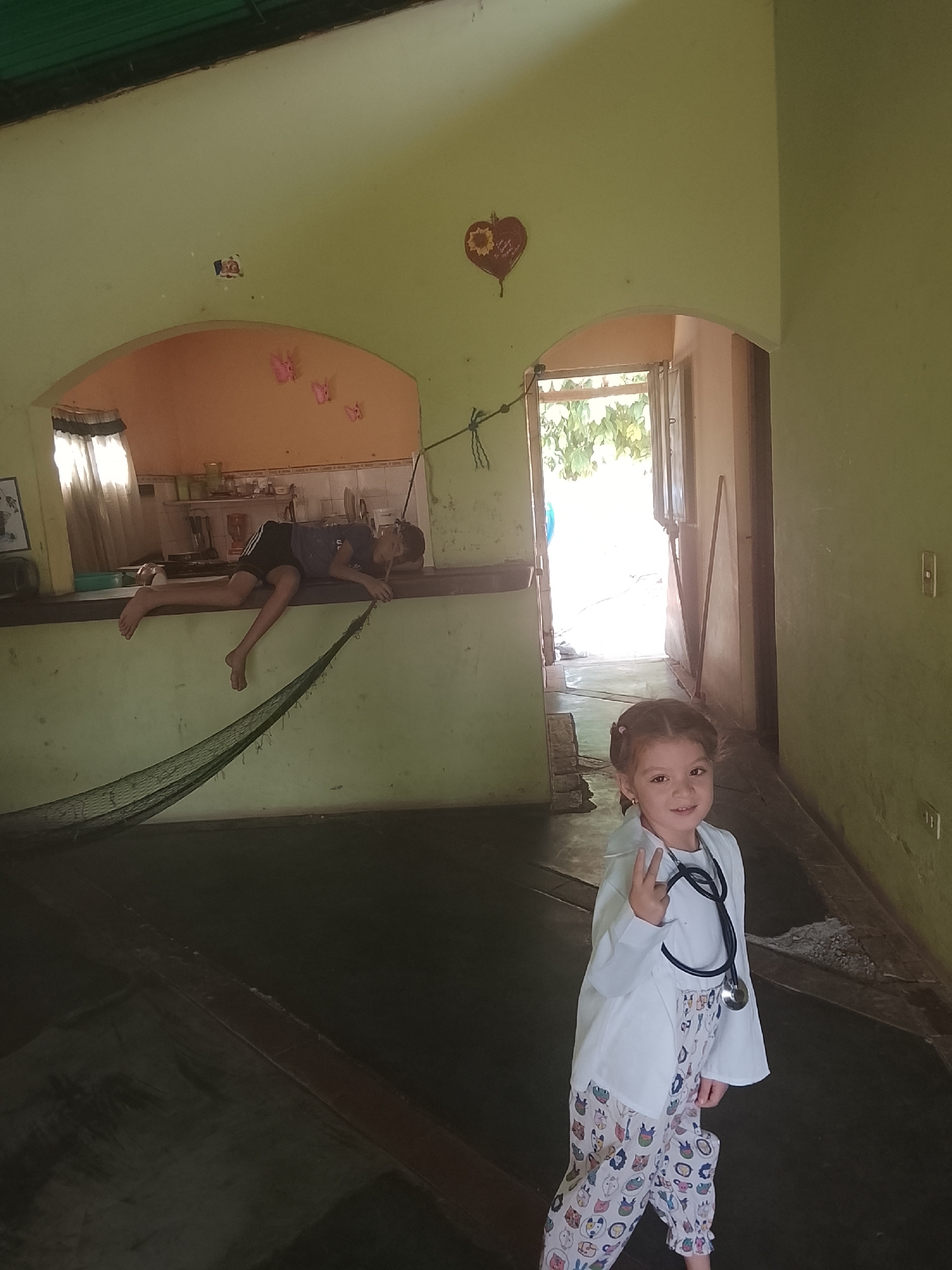 A full shot captures the interior of a residence in Altagracia, Venezuela. The walls are painted in a pale green hue, and the floor is dark. An opening leads into what appears to be a kitchen area. A boy is lying down on top of a wall that is a part of the kitchen wall looking over a net like object in the main room.
A young girl stands in the foreground, dressed in pajamas and a white lab coat, with a stethoscope around her neck. She holds up two fingers in a "peace" sign.
An open doorway allows a view to the outdoors with green foliage.