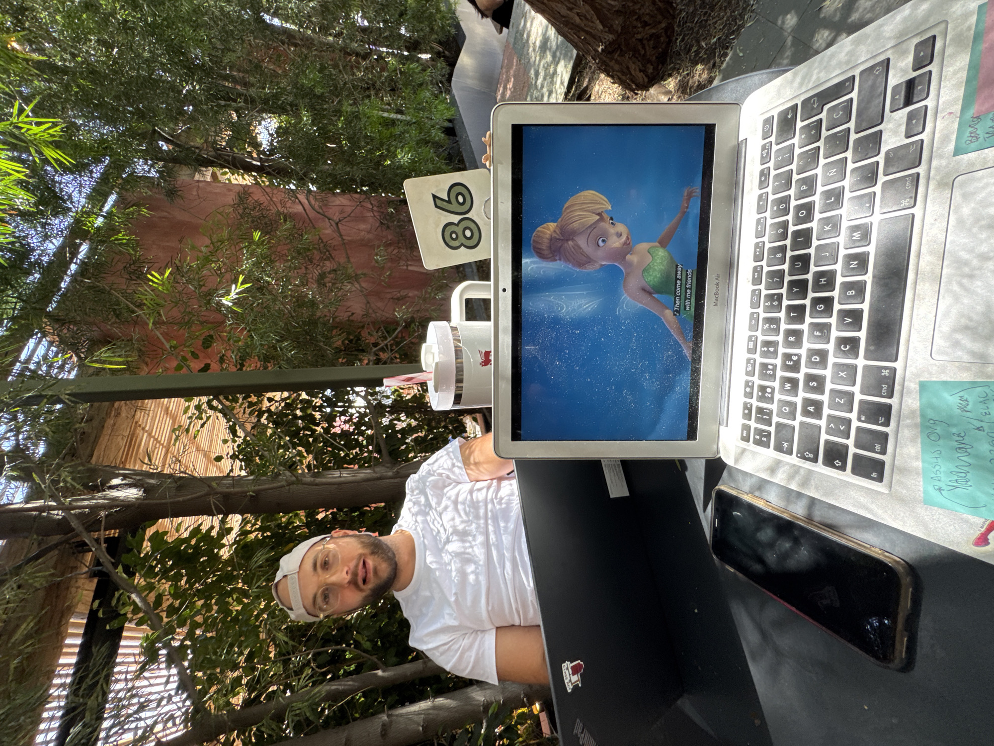 This image, rotated 90 degrees counter-clockwise, depicts a man at an outdoor table in a cafe or restaurant setting in West Hollywood, United States of America. Bright sunlight suggests it is daytime.
The subject is a man wearing a white t-shirt, clear-rimmed glasses, and a light-colored baseball cap worn backward. He is seated at a dark table, looking directly at the camera with a neutral expression.
On the table, a silver MacBook Air is open, displaying an animated scene featuring Tinkerbell, with the subtitle "Then come away with me friends" visible on the screen. To its right, a dark-colored laptop, possibly a ThinkPad (based on the visible "Pad" text and a "CLASS ACT" sticker, and a partial "MARVE" sticker), is closed. A black smartphone with a gold-colored case lies face down next to the laptops. A white table number "86" is positioned near the MacBook Air. Two stacked white beverage cups, one with a red logo, are also on the table. A light blue sticky note attached to the MacBook's chassis contains handwritten text including "ASSIS. Olg", "Yelamane", "ELAC", and "2260".
The background features abundant green foliage and trees, indicating an outdoor dining area, with glimpses of a reddish-brown building or wall through the leaves. The man appears to be relaxing or working in a casual, sunny outdoor environment.