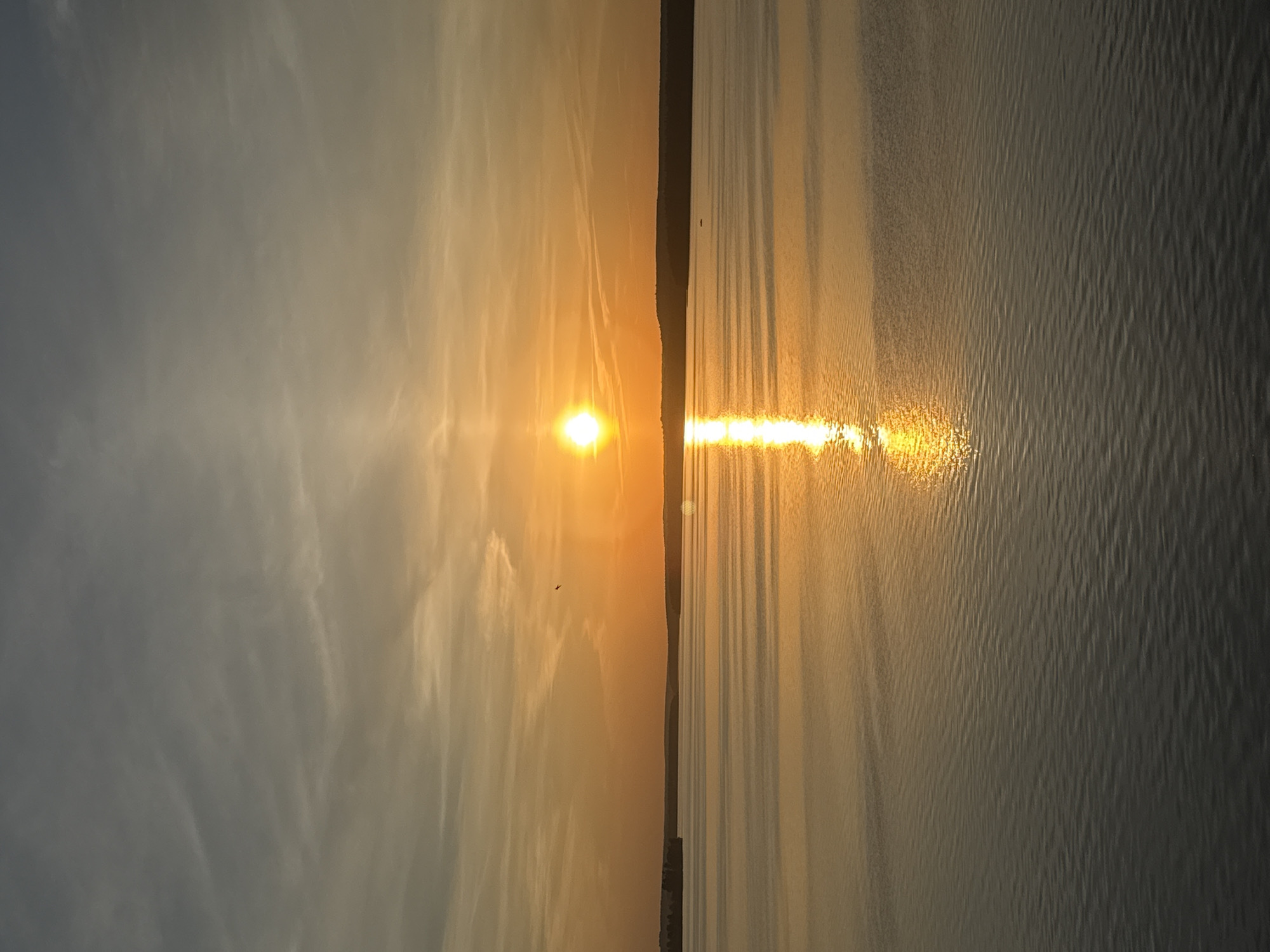 The image depicts a sunrise or sunset scene over a body of water. The sun is the central focus, with its light reflecting brightly on the water's surface. The sky transitions from a light orange near the sun to a dimmer grey/blue further away. There is a dark landmass or coastline visible between the sky and water.
While Votkinsk, Russia is known for its reservoir on the Kama River, this image does not allow definitive confirmation of the location.