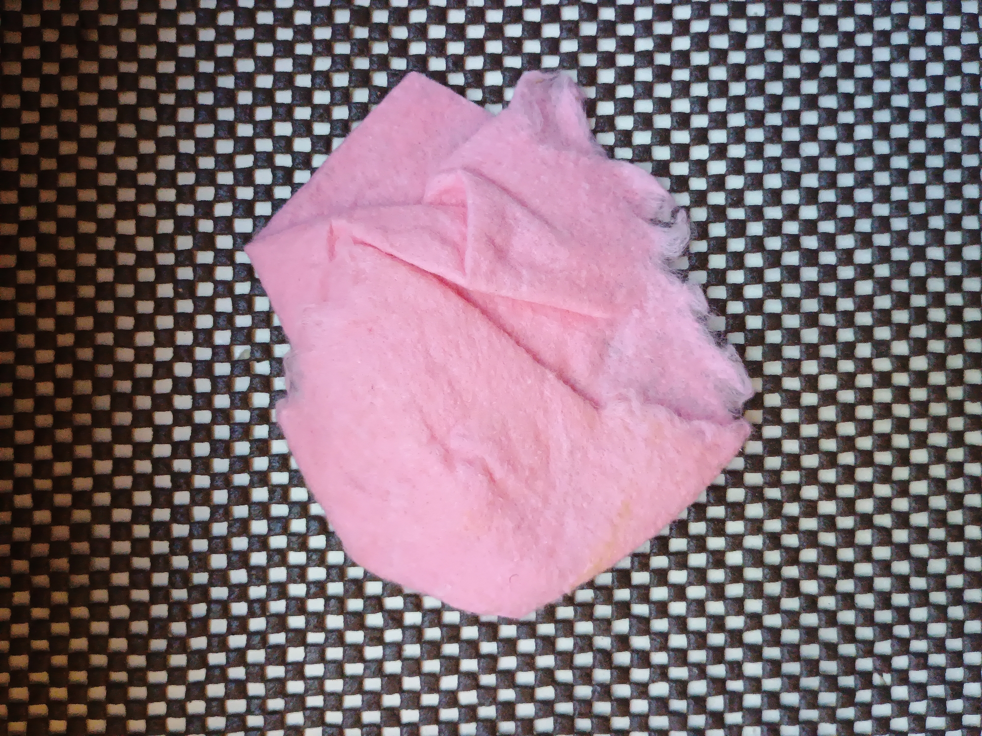 A close-up, overhead shot shows a crumpled piece of pink fabric or tissue paper on a textured surface. The surface is a checkered pattern of dark brown and white squares. The pink material is bunched up, with visible folds and a slightly frayed edge on the right side. The fabric appears soft and possibly fuzzy. The lighting suggests an indoor setting, with no harsh shadows. There is no visible text. The image does not depict any people or a specific event. The location context of Zaporizhzhia, Ukraine, is provided but not visually discernible from the image itself.