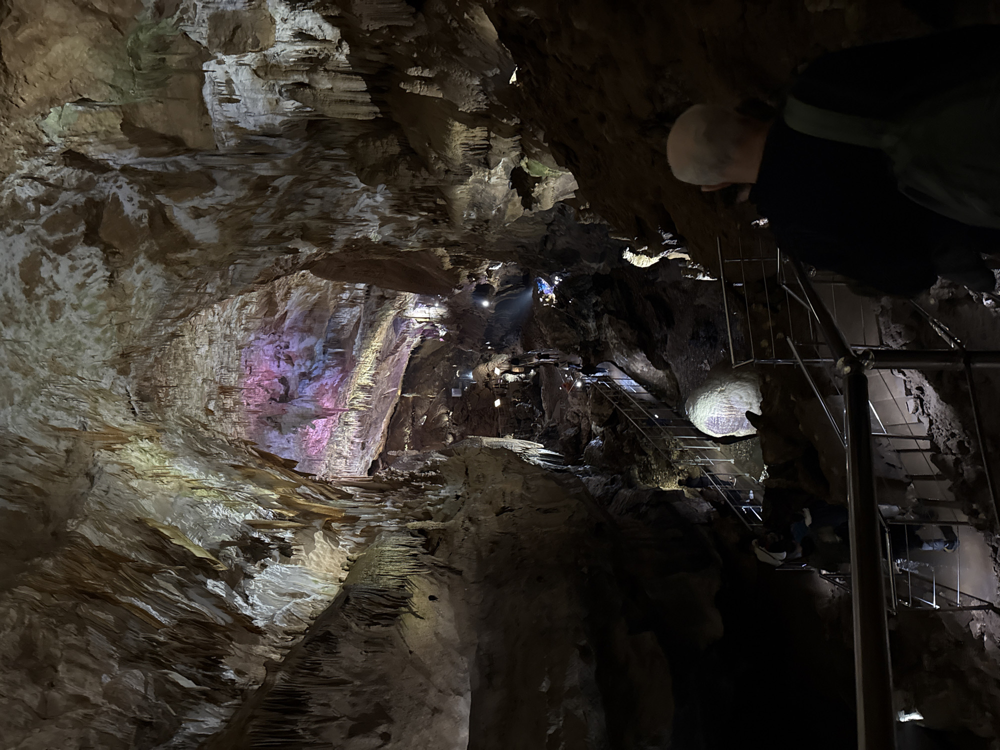 The image depicts the Prometheus Cave in Tskaltubo, Georgia. The cave features illuminated geological formations, including stalactites and stalagmites. A metal staircase with railings provides access for visitors. Multiple people are observed on the staircase and in the cave interior. A man is partially visible in the upper right corner. The lighting in the cave is artificial, with pink, yellow, and white hues highlighting the rock textures.