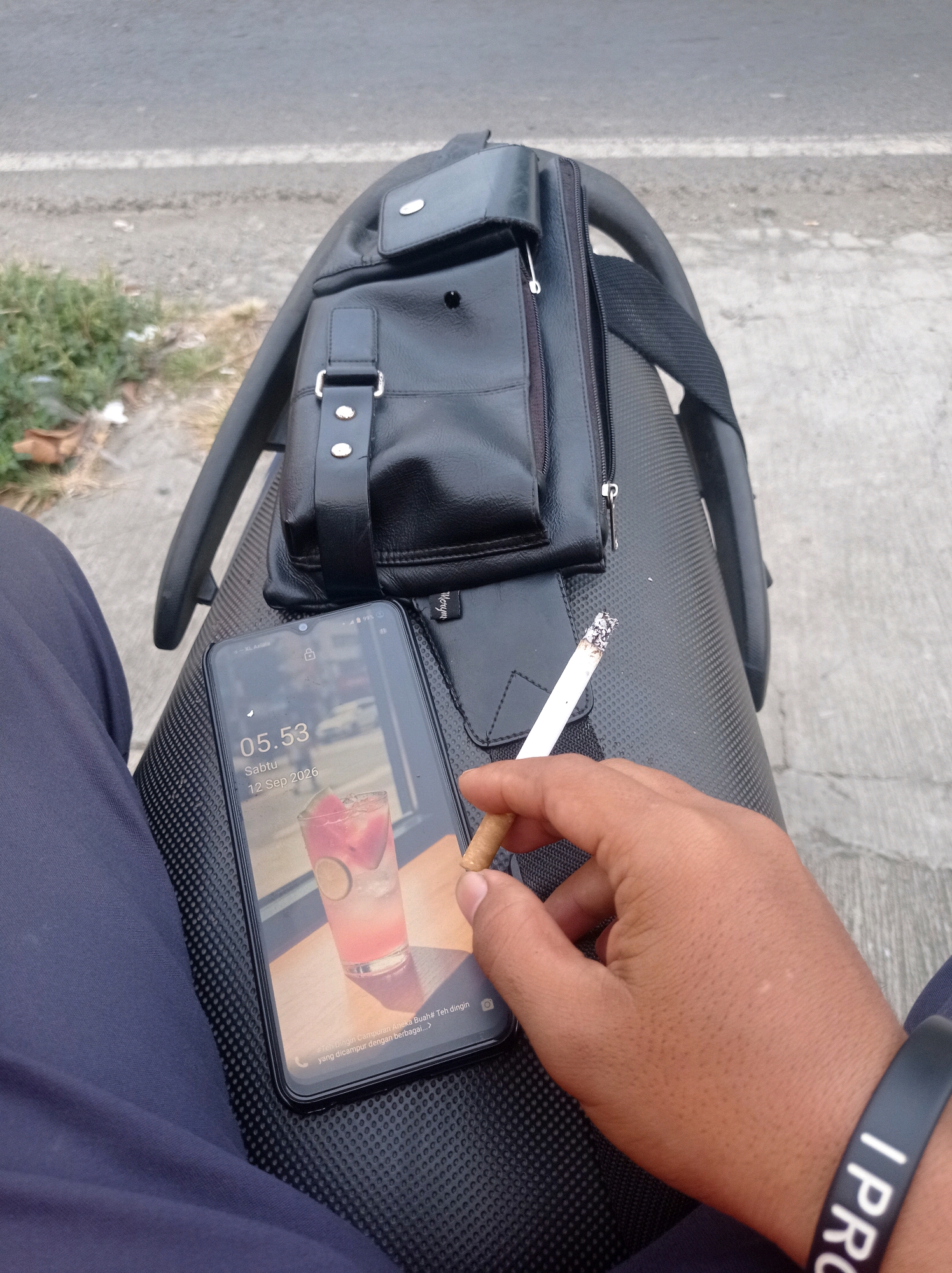 The image shows a close-up view of a person's hand holding a lit cigarette. The cigarette is positioned over a dark, textured surface, likely a motorcycle seat. To the left of the hand is a smartphone displaying the time "05:53" and the date "Sabtu 12 Sep 2026". The phone also shows a lock screen image of a pink drink with ice and fruit. Behind the phone and cigarette is a black leather bag with a buckle closure. The background suggests an outdoor setting, with a paved road and some greenery visible. The lighting indicates it is likely daytime, possibly early morning. The image does not provide specific details about the location within Jetis, Indonesia, beyond the implied urban or suburban environment suggested by the road.