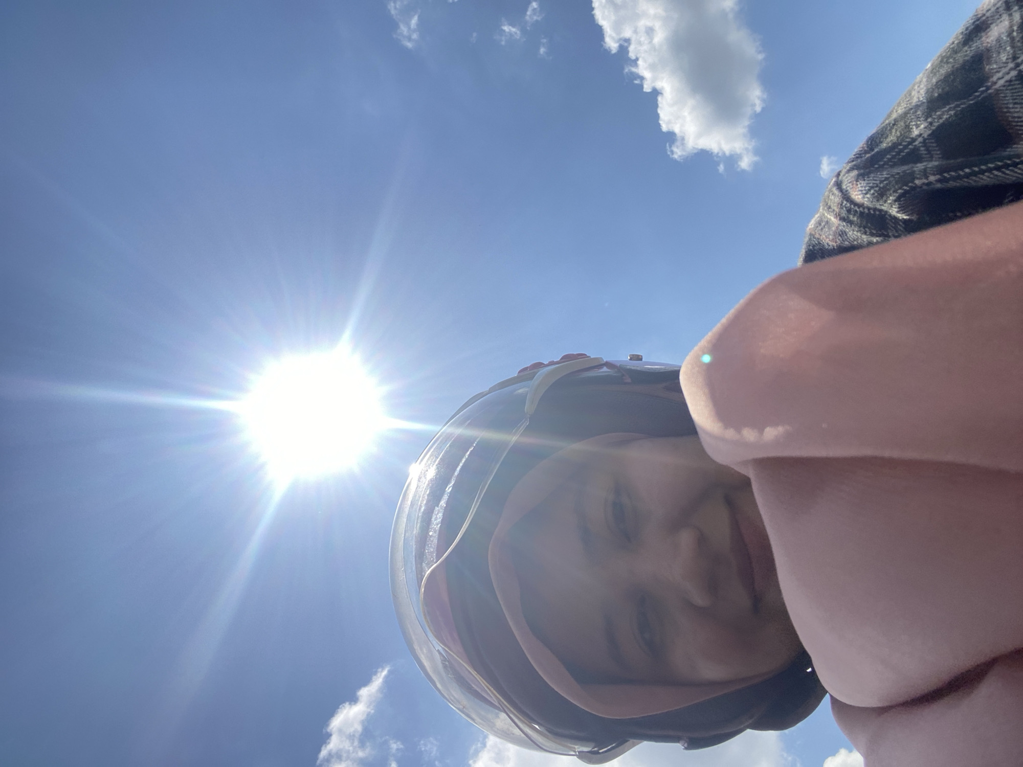A woman wearing a pink hijab and a helmet smiles at the camera in a selfie. She is wearing a plaid shirt. The sun is bright and high in the sky, with visible sun flares. There are a few white clouds in the blue sky. The woman is holding a pink scarf or blanket in front of her. The overall scene appears to be outdoors during the day, possibly on a motorcycle due to the helmet. The location is the city of Banjarmasin, Indonesia.