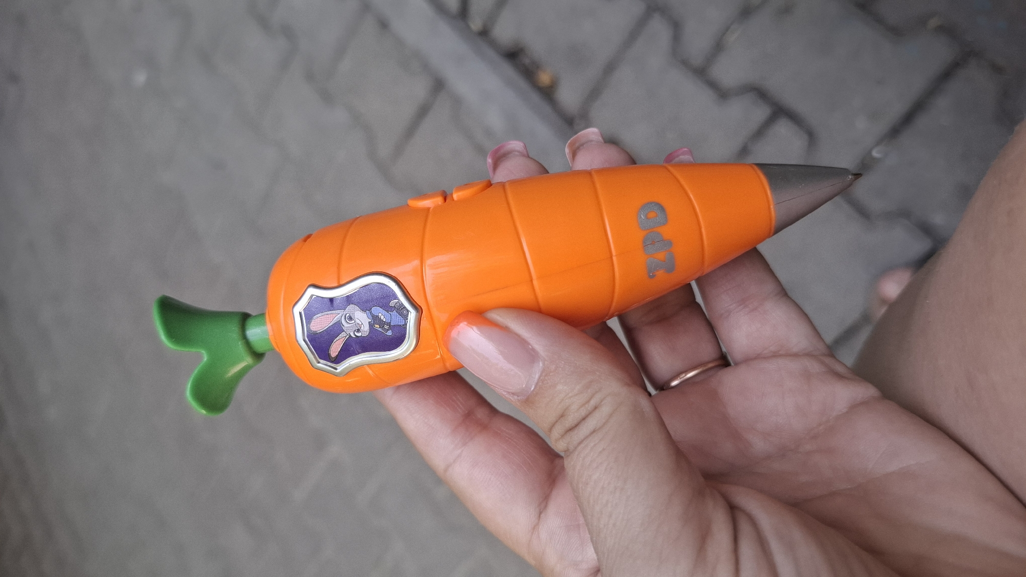 A hand with manicured nails, painted with a gradient of pink to orange, holds an orange, carrot-shaped object. The object has a green leaf sprout at its top and a silver-colored tip. A purple badge with a cartoon rabbit wearing a blue police uniform is affixed to the object. The letters "ZPD" are embossed in silver on the orange surface below the badge. The background is a paved surface composed of gray interlocking bricks. The image appears to have been taken outdoors in the city of Shymkent, Kazakhstan.