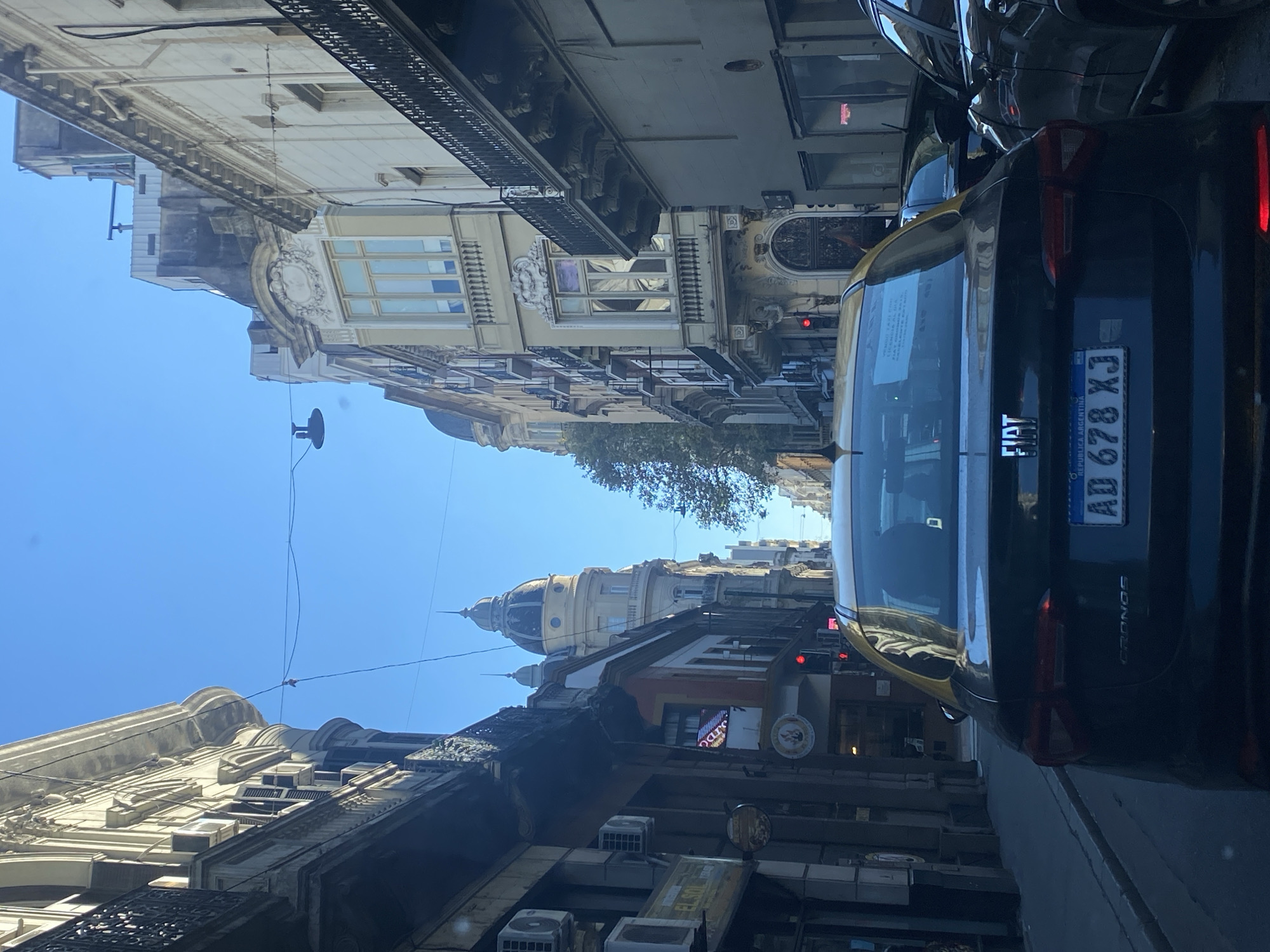 The image shows a street scene in Buenos Aires, Argentina. The camera is positioned inside a vehicle, looking out towards the street. A black Fiat taxi is in the foreground, with its license plate "AD 678 XJ" visible. The street is lined with buildings of various architectural styles, including ornate facades with balconies. There is a building with a dome. There are signs and shopfronts along the street. The sky is blue and clear. The traffic signal is red.