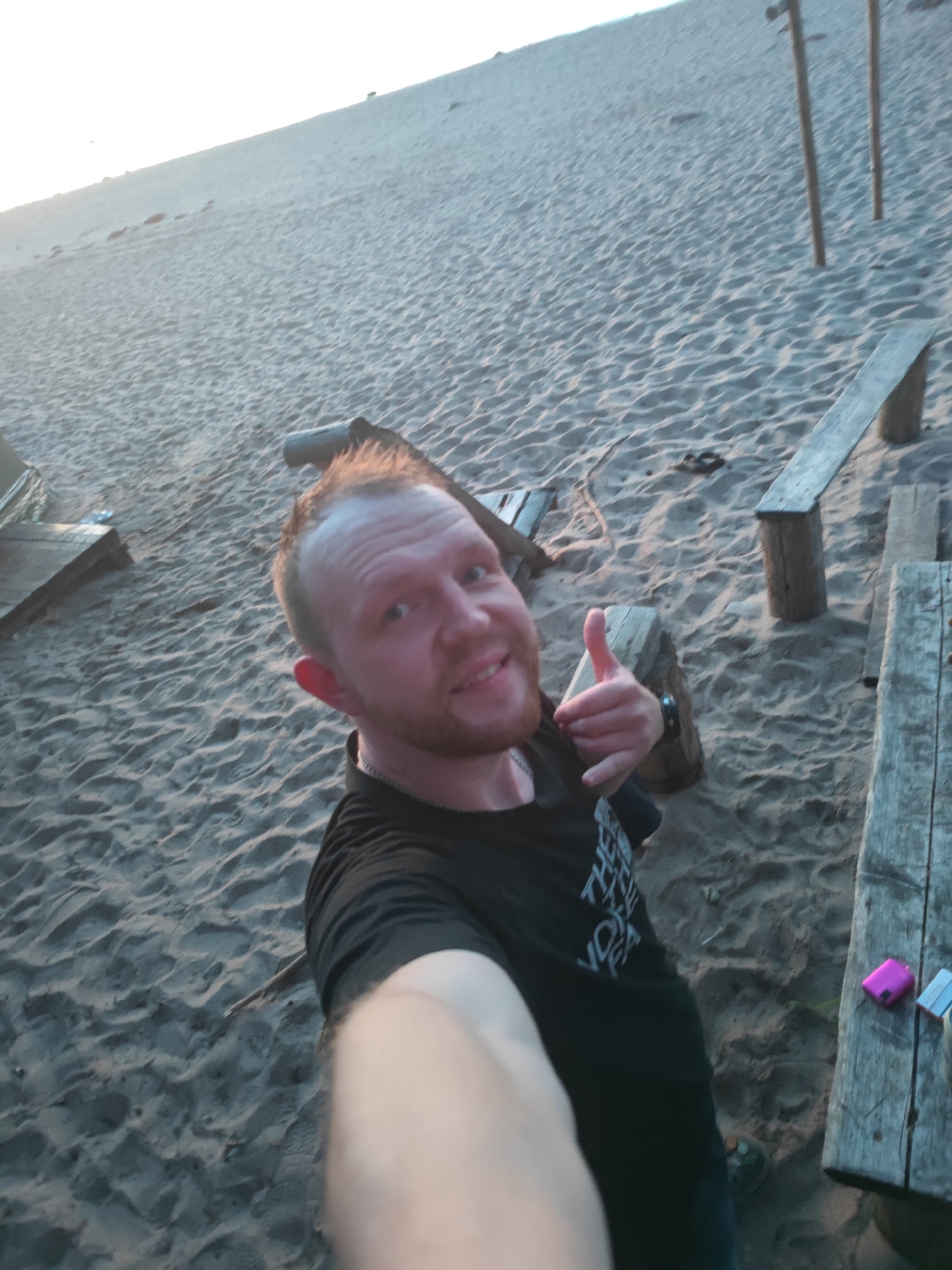 A man with short brown hair and a beard is taking a selfie on a sandy beach. He is wearing a black t-shirt and has his left arm extended to take the photo. His right thumb is raised in a "thumbs up" gesture. The beach is covered in sand dunes and there are wooden benches and structures scattered around. The sky in the background is bright and overcast, suggesting it is either early morning or late evening. There are no other people visible in the image.