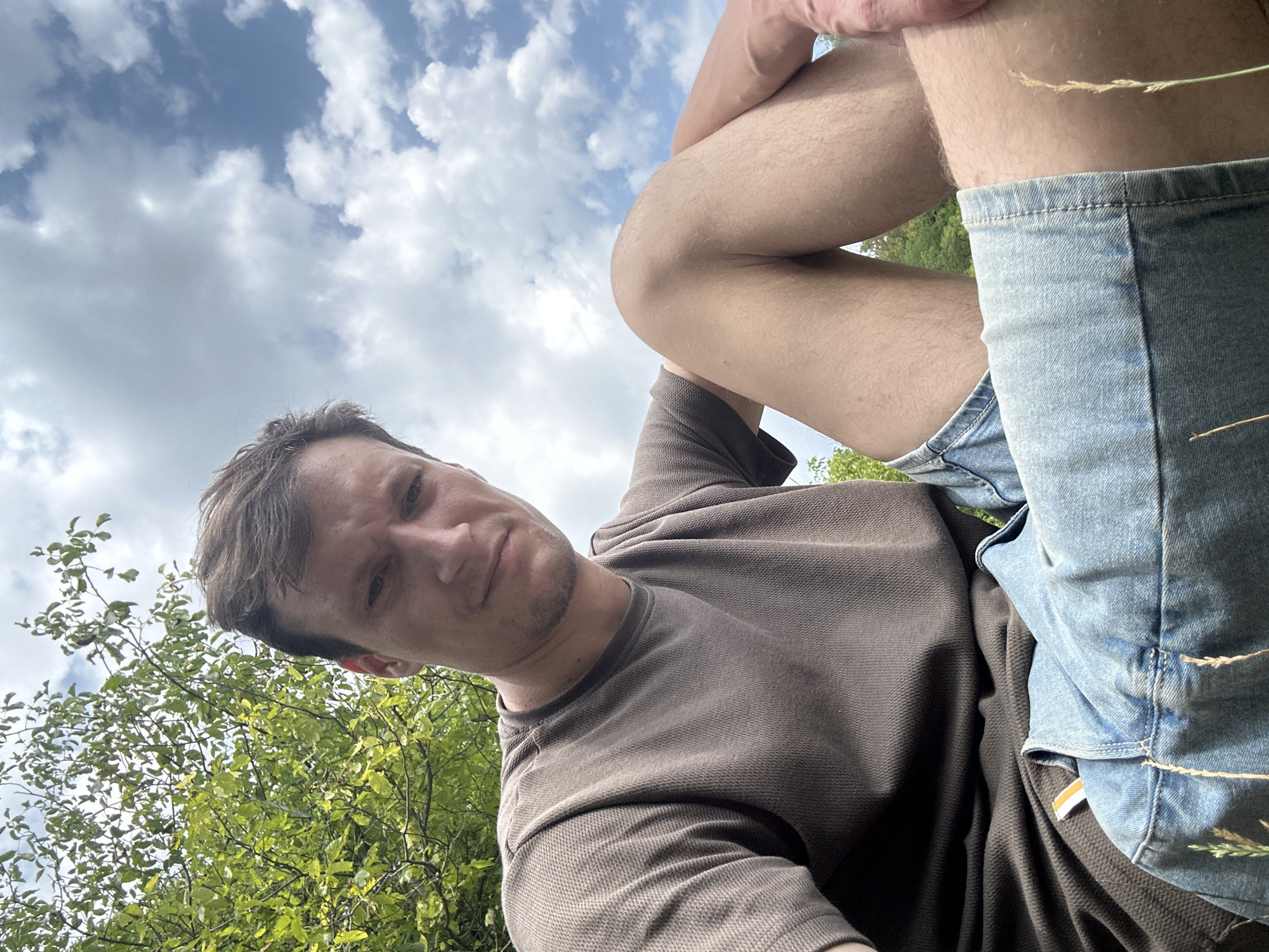A low-angle, over-the-shoulder selfie captures a man sitting outdoors. He is wearing a brown textured t-shirt and denim shorts. His face is turned towards the camera, with a slight smile. The background features lush green trees and a cloudy blue sky. The location is identified as Makiv, Ukraine.