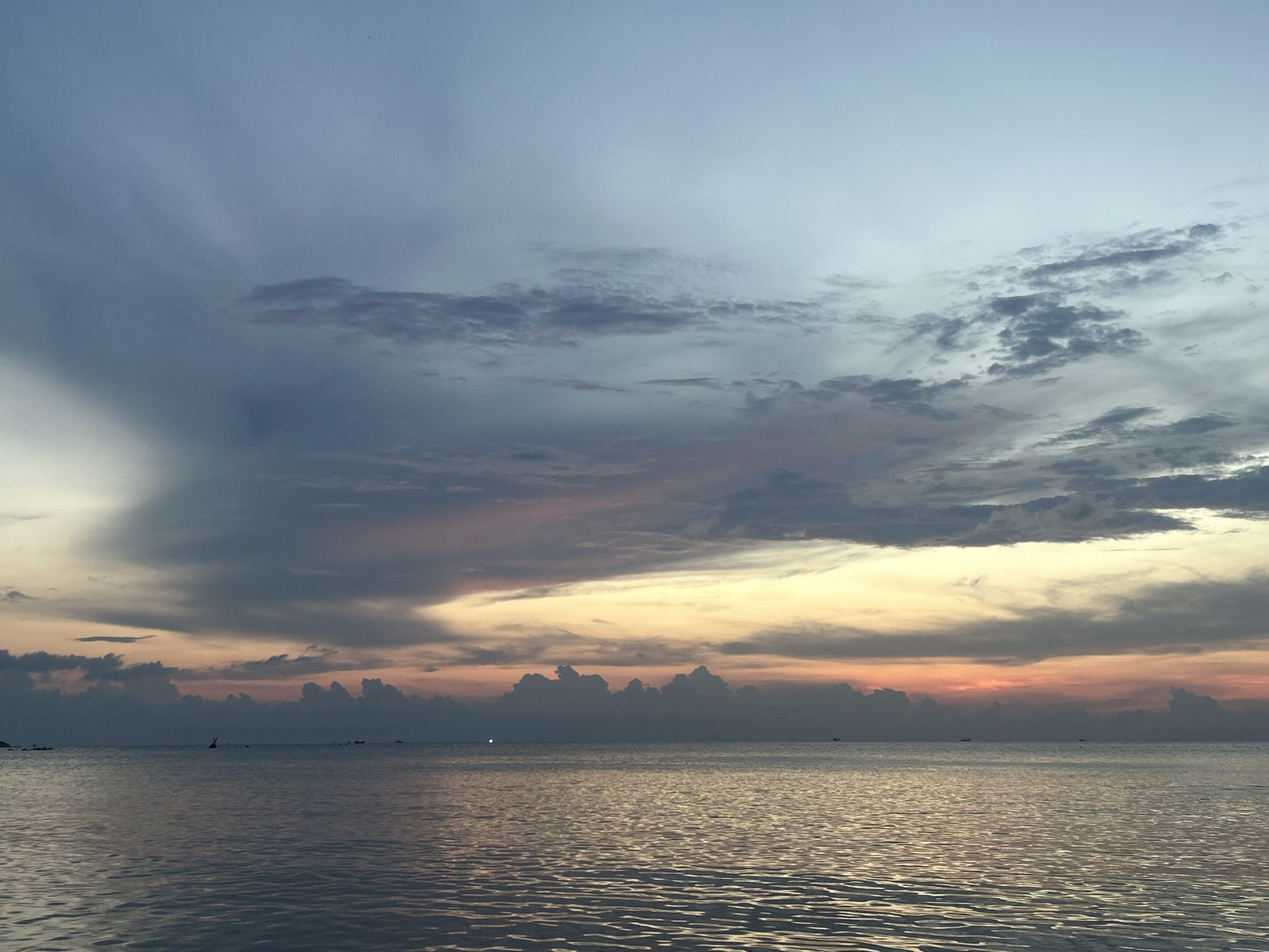 The image depicts a seascape at sunset in Baan Wang Ta Kien, Thailand. The sky is filled with layers of clouds, transitioning from shades of blue at the top to yellow, orange, and grey hues closer to the horizon. A band of dark, cumulonimbus clouds sits near the water's edge. The sea reflects the colors of the sky, with ripples and waves creating a shimmering effect. Small boats are visible on the horizon.