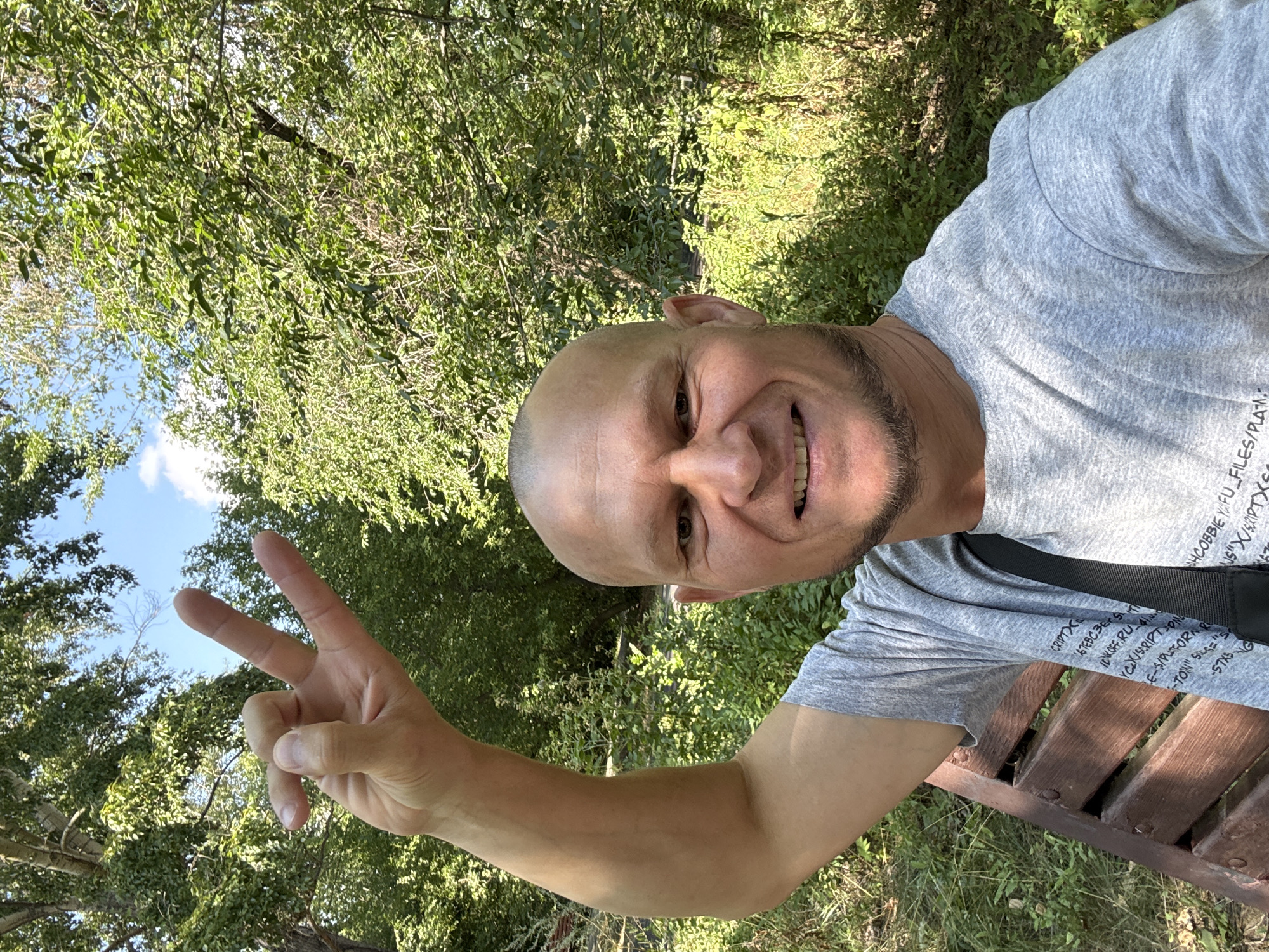 A bald man in a gray t-shirt smiles for a selfie in a park, raising his left hand with his thumb and index finger touching, forming a circle, and his middle and ring fingers extended upwards in a 'V' shape, against a backdrop of lush green trees and a blue sky with a few clouds. The man is seated on a wooden bench, with a black strap from a bag visible over his shoulder. The setting appears to be a natural, outdoor environment, possibly a public park within the city of Oskemen, Kazakhstan, characterized by dense foliage and trees.