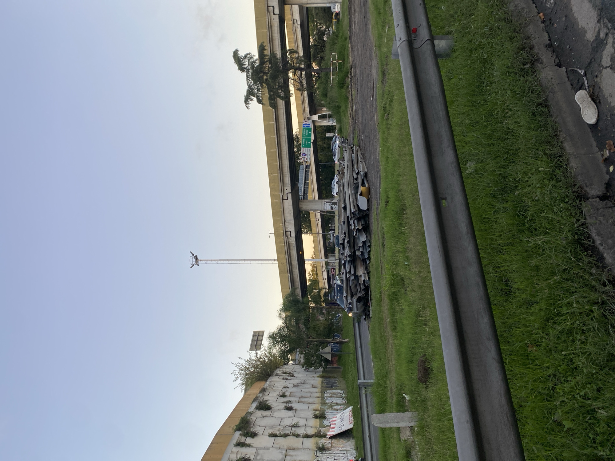 The image depicts an outdoor scene in what appears to be Buenos Aires, Argentina. A segment of an elevated highway structure dominates the background. Underneath it, a roadside scene unfolds with patches of green grass. A metal guardrail runs along the edge of the road, with a gray road surface. Scattered piles of metal tubes or pipes can be seen. On the right edge of the frame, there's a discarded white shoe. Beyond the metal piles, a concrete retaining wall with vegetation growing out of the cracks is visible, along with remnants of graffiti or discarded materials. A tall metal tower stands near the highway structure. The sky is light blue and the overall lighting suggests either early morning or late afternoon.