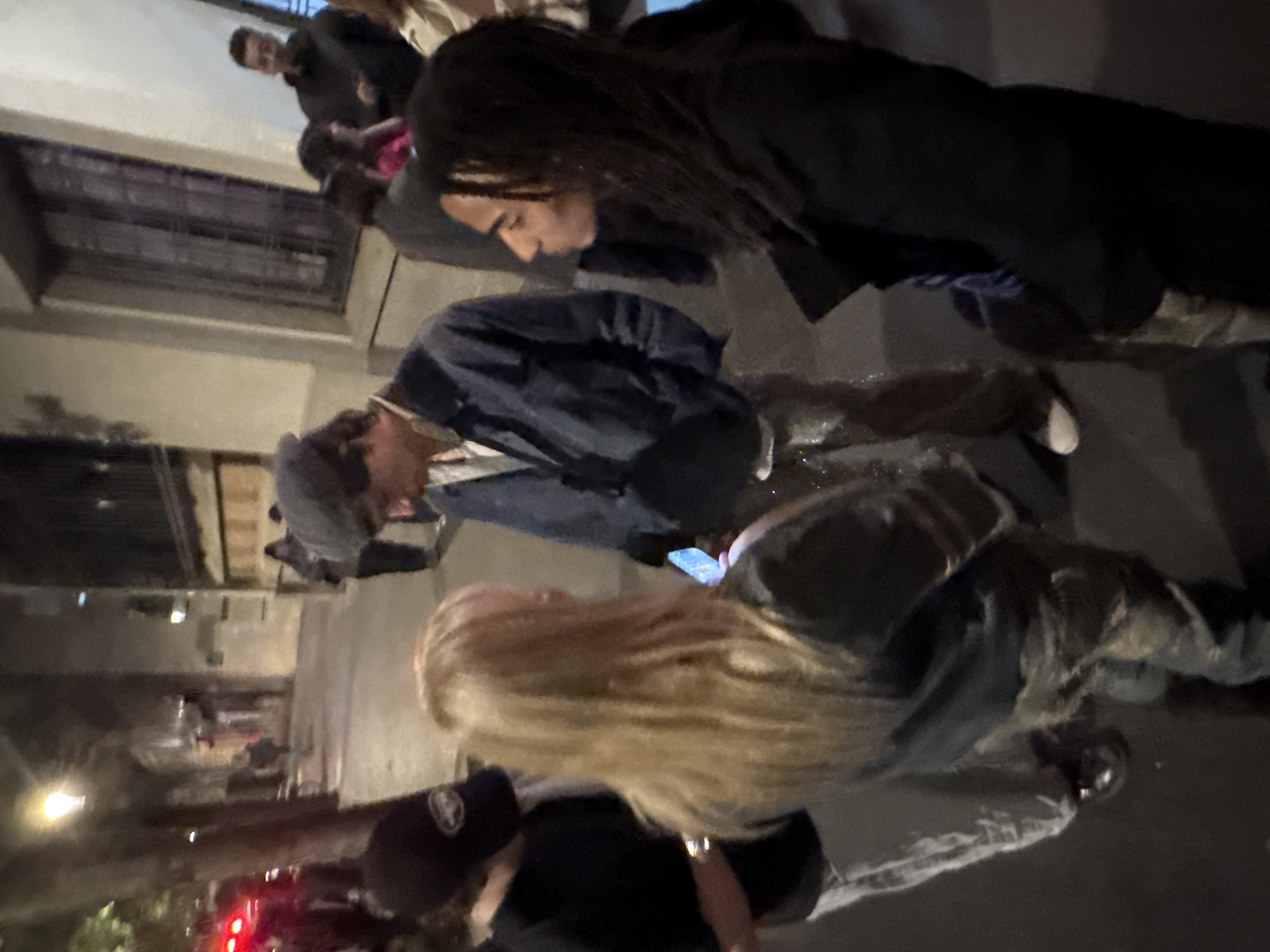 This image depicts a nighttime urban scene, likely a street or public area in Paris, France. Several individuals are visible, illuminated by artificial light sources.
In the foreground, a person with long, blonde hair is partially shown, wearing a dark jacket and looking downwards. Beside them, another person in a dark cap and dark attire is also present. In the mid-ground, a person with long, dark hair, possibly dreadlocks or braids, is captured in profile, wearing a dark jacket and also looking downwards. Next to this individual, another person in a dark blue jacket and a cap is engrossed in a smartphone, which emits a distinct blue glow from its screen.
Further back, additional people are faintly visible, including one wearing a black jacket over a pink top, and another person with short dark hair. The overall lighting is dim, characteristic of night, with a bright streetlight or lamp visible in the background and red lights, possibly from a vehicle or traffic signal, in the lower left. Structures resembling buildings and metal grates can be seen in the background, reinforcing the urban setting. The individuals appear to be standing or loitering, with some focused on their phones or something on the ground. No specific event or strong emotions are discernible, and no text is clearly visible.