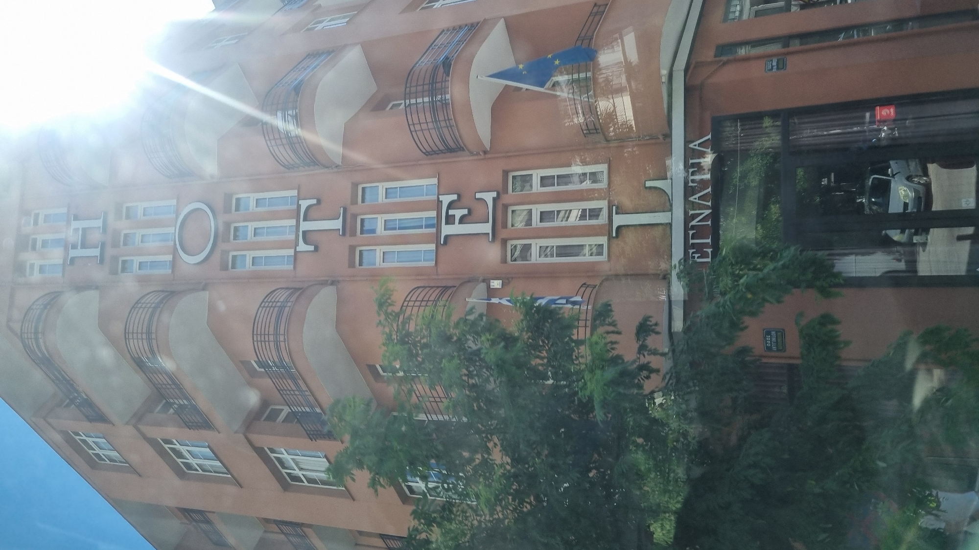 The image shows the exterior of a building, likely a hotel, in Thessaloniki, Greece. The word "HOTEL" is prominently displayed vertically on the facade in large, silver block letters. Below the hotel sign, the word "EGNATIA" is visible, which is the name of the street or a business located there. The building has a reddish-orange facade and features multiple balconies with black railings. There are flags visible on the balconies, one appears to be the Greek flag and another is the European Union flag. A large tree with green foliage partially obscures the left side of the building. The sky is visible at the top, and there is a bright glare from the sun in the upper right portion of the image. The ground floor of the building has large glass doors, through which a car is visible inside. The overall scene depicts a street view of a building in an urban environment.